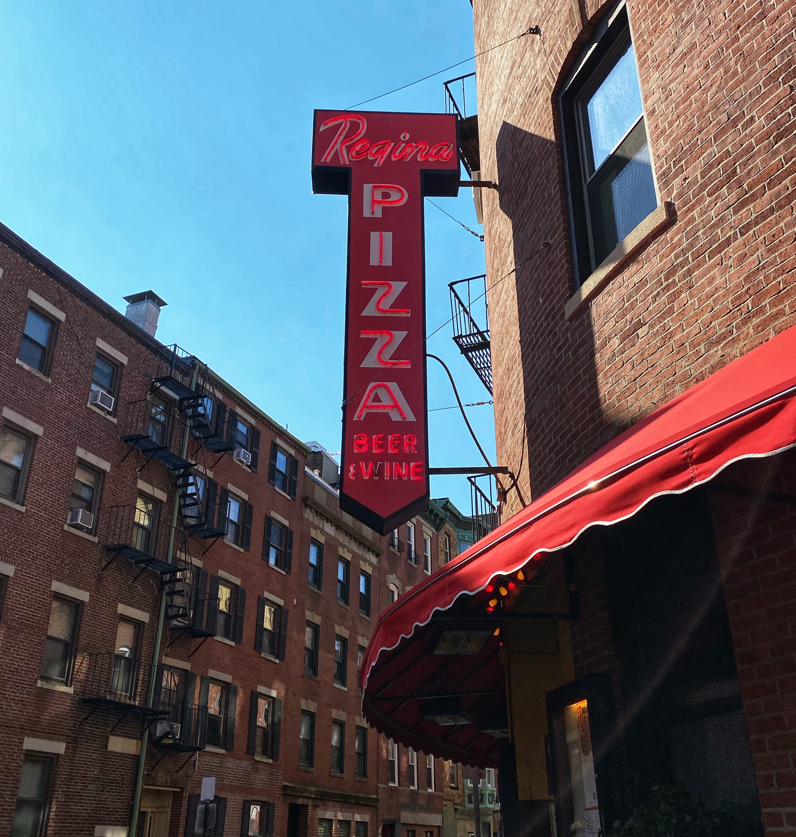 A sign for Regina Pizzeria hangs on the corner of a building with a red awning beneath the neon sign.