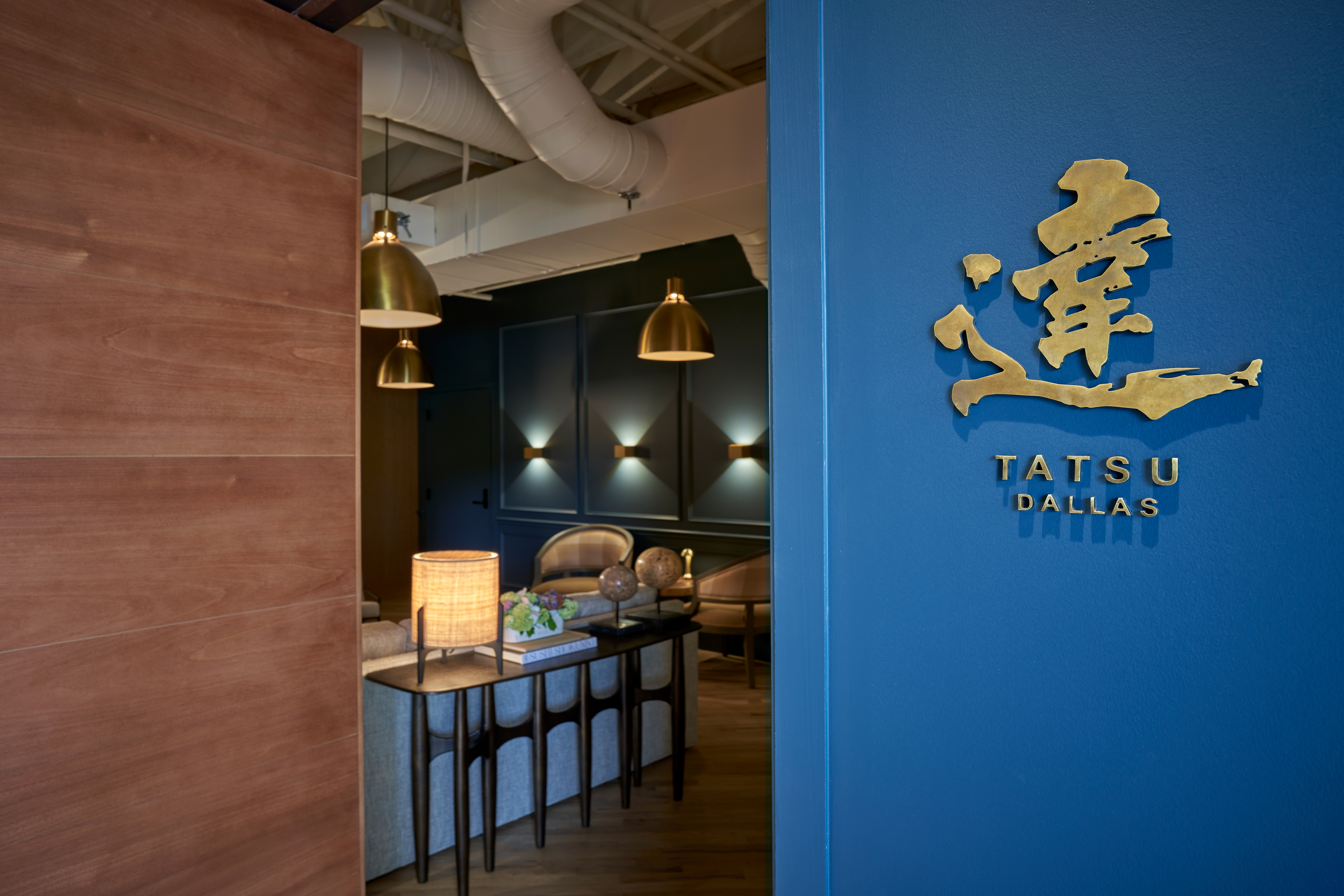 Between a blue door with the words “Tastu Dallas” and a gold design, and a wooden wall, peeks out a glimpse into a small dining room where sushi is served.