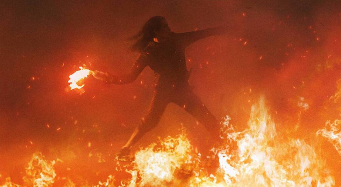 A man with long hair throws a molotov cocktail while enveloped by fire in Athena