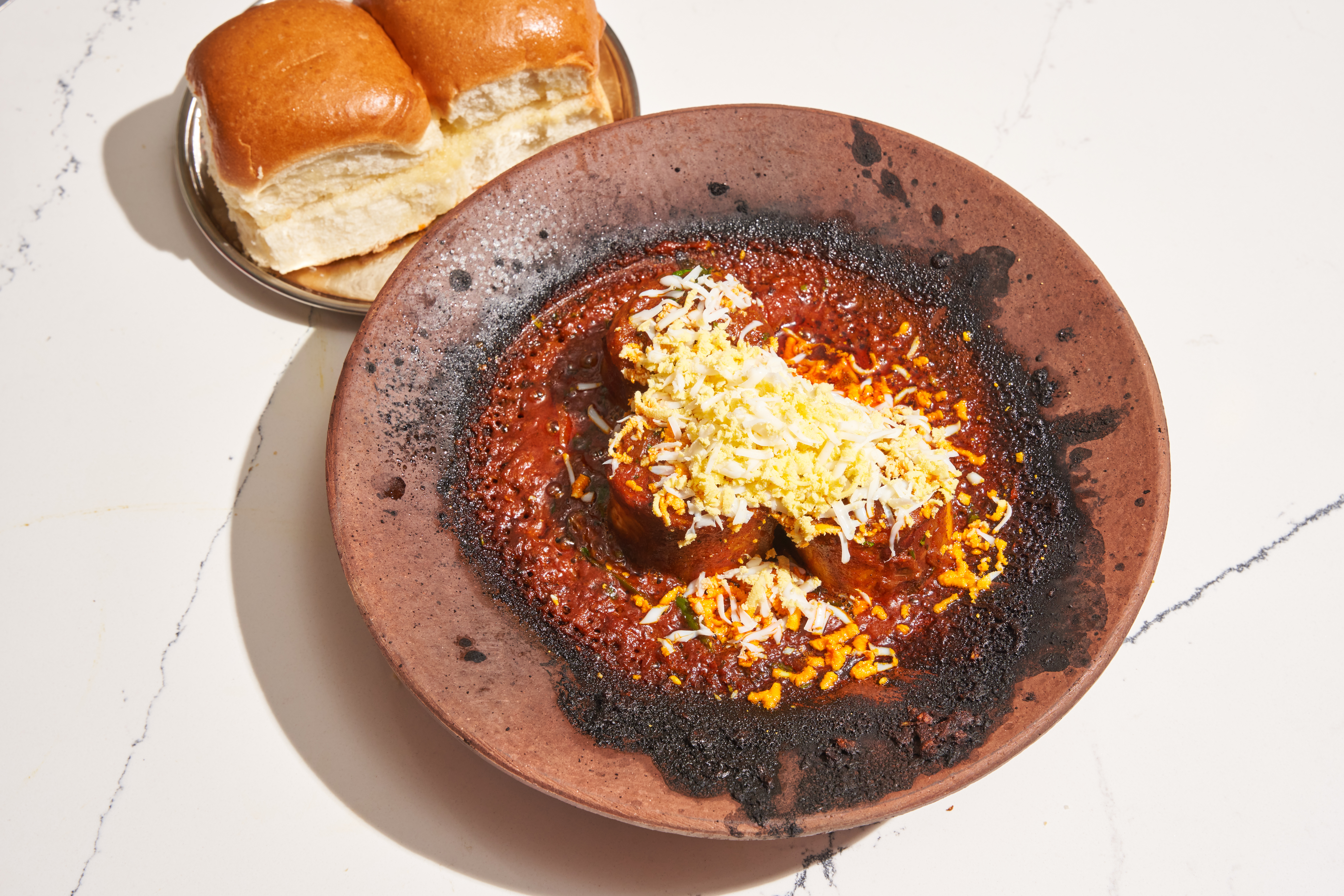 A wide-mouthed clay bowl of beef and red sauce with two golden buns on a small plate off to the side.