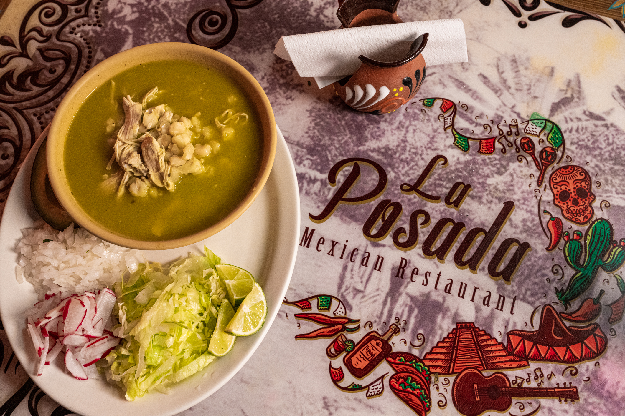 A bowl of green pozole with garnishes and the cover of a menu that says La Posada in Detroit, Michigan.