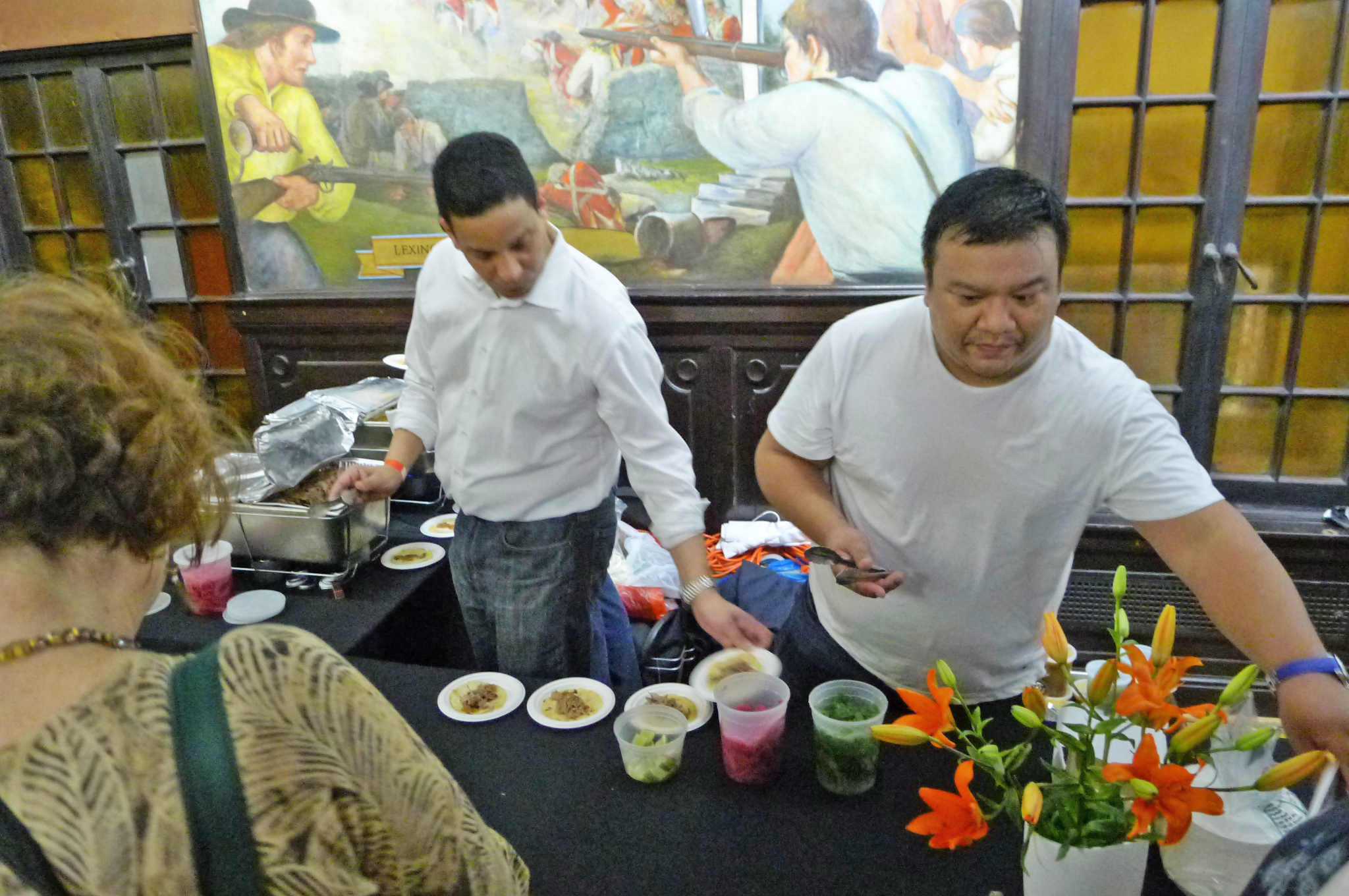 Two chefs work behind a table lined with small plates as they greet diners at a food festival.
