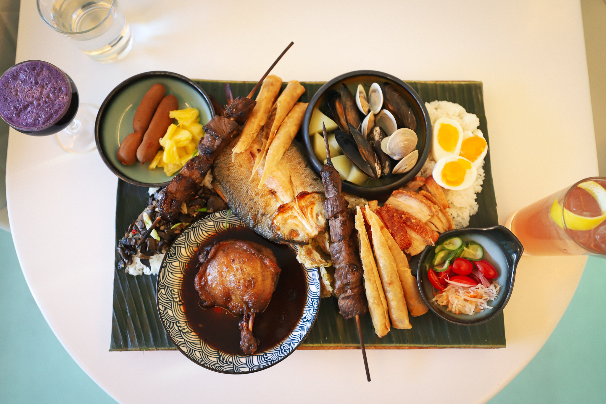 A single-person kamayan feast with food laid out on a green banana leaf the size of a placemat. Drinks are off to the sides.