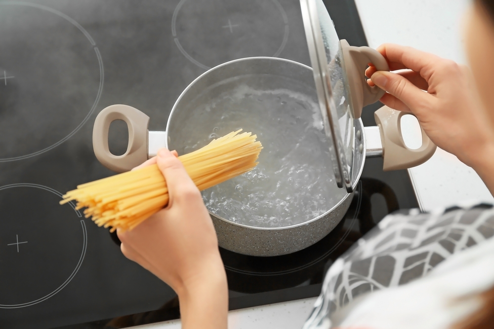 A woman stands over a pot of boiling water, holding a handful of spaghetti in her left hand and the lid to the pot in the right