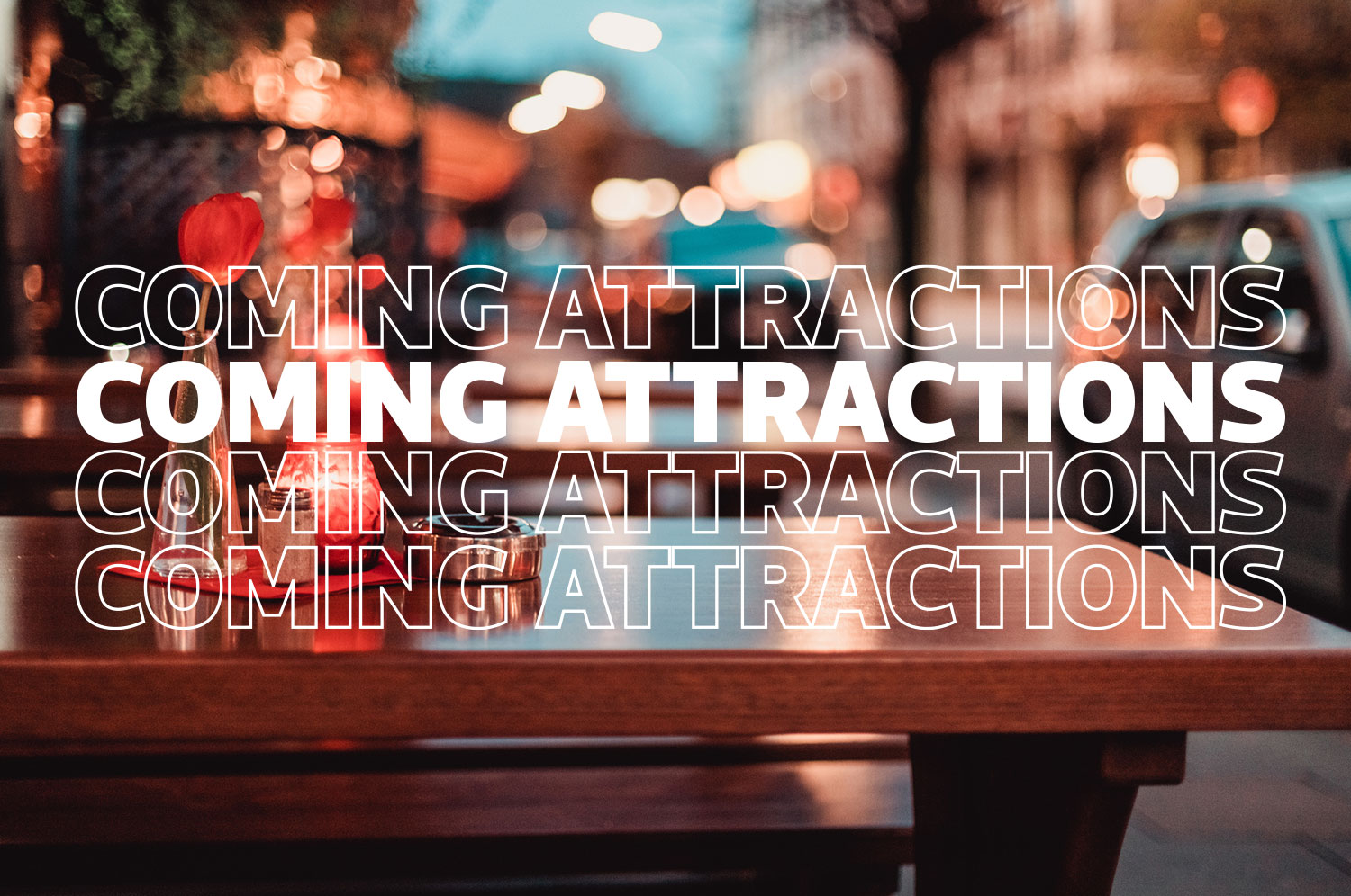 City lights shine in the evening background and a wooden picnic style table sits in the foreground. White text ripples down the page reading, “Coming Attractions.”