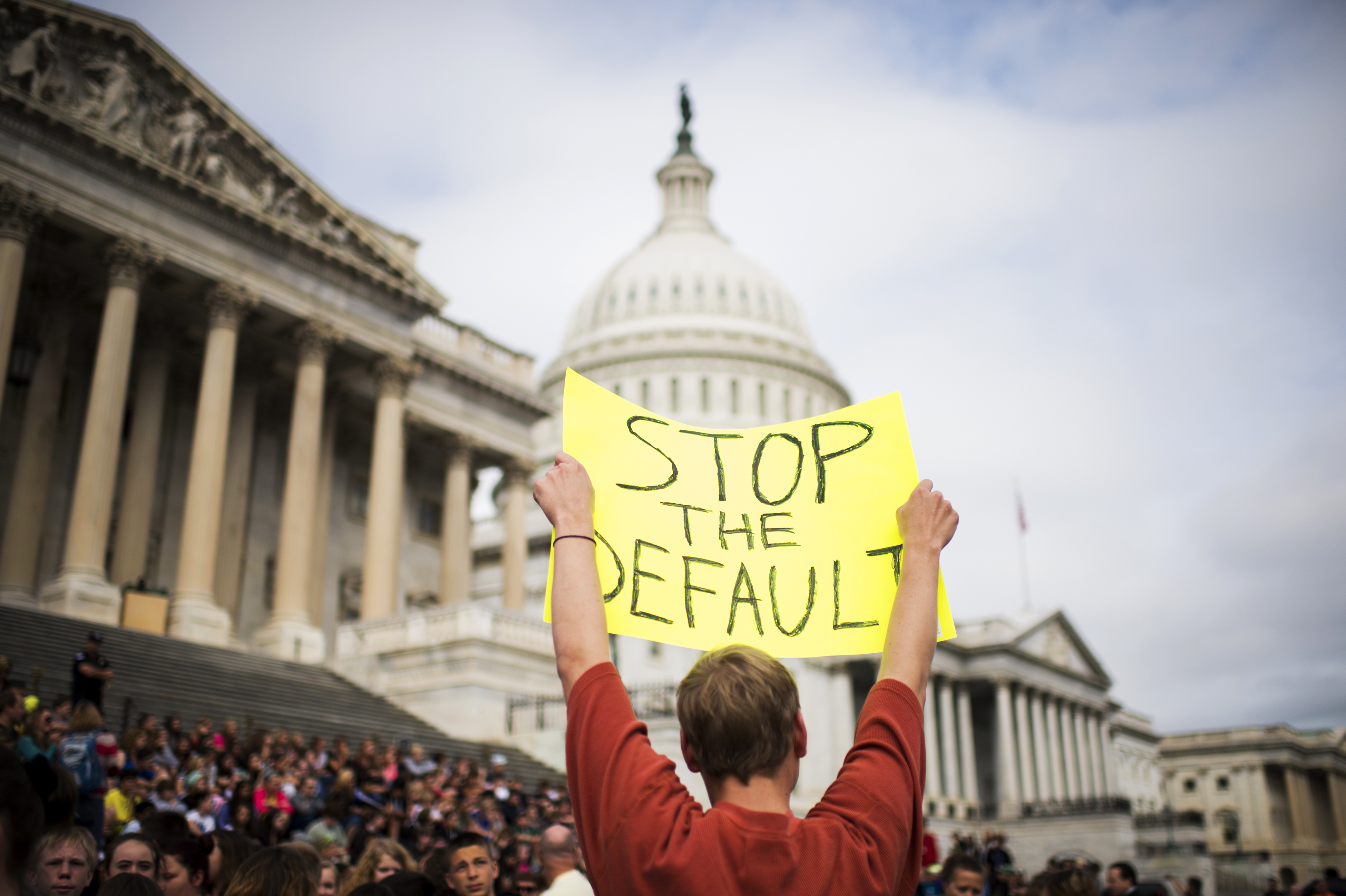 A protester at the US Capitol building holds a hand-lettered sign over their head that reads “Stop the default.” 