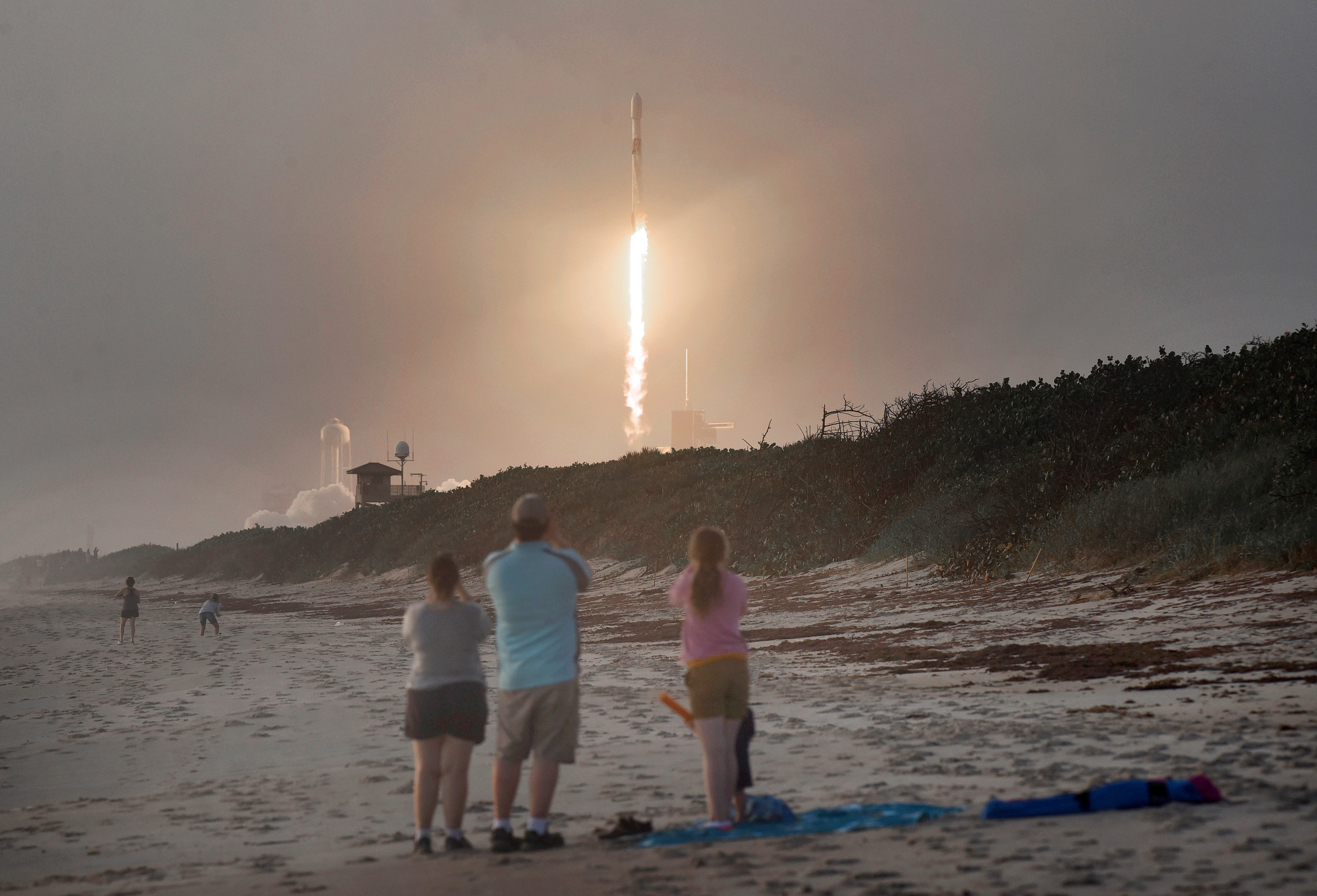 Three people stand on a beach watching a SpaceX rocket climb into the sky.