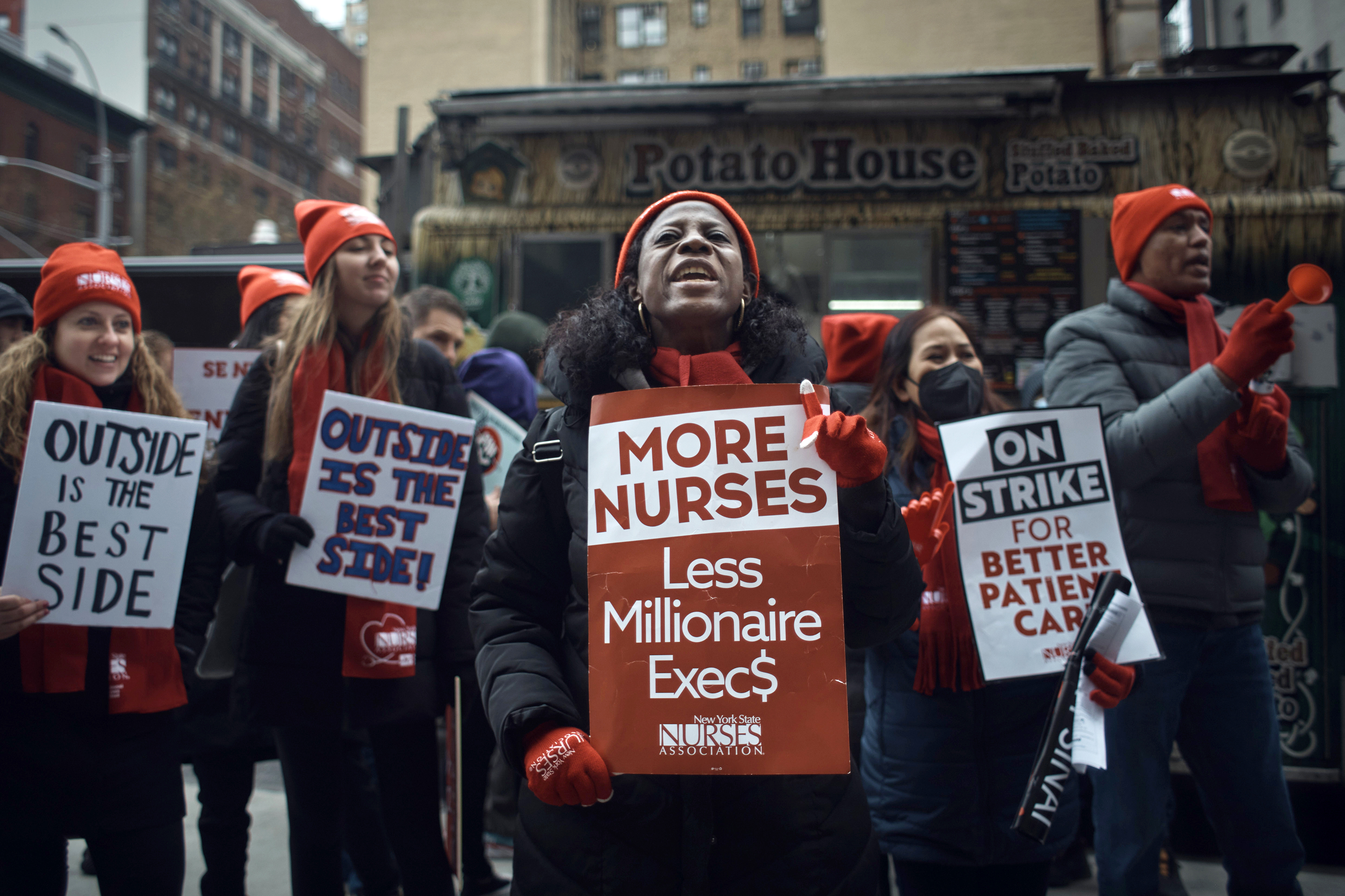 Protesters outside a hospital hold signs that read, “More nurses, less millionaire execs,” “Outside is the best side,” and, “On strike for better patient care.”