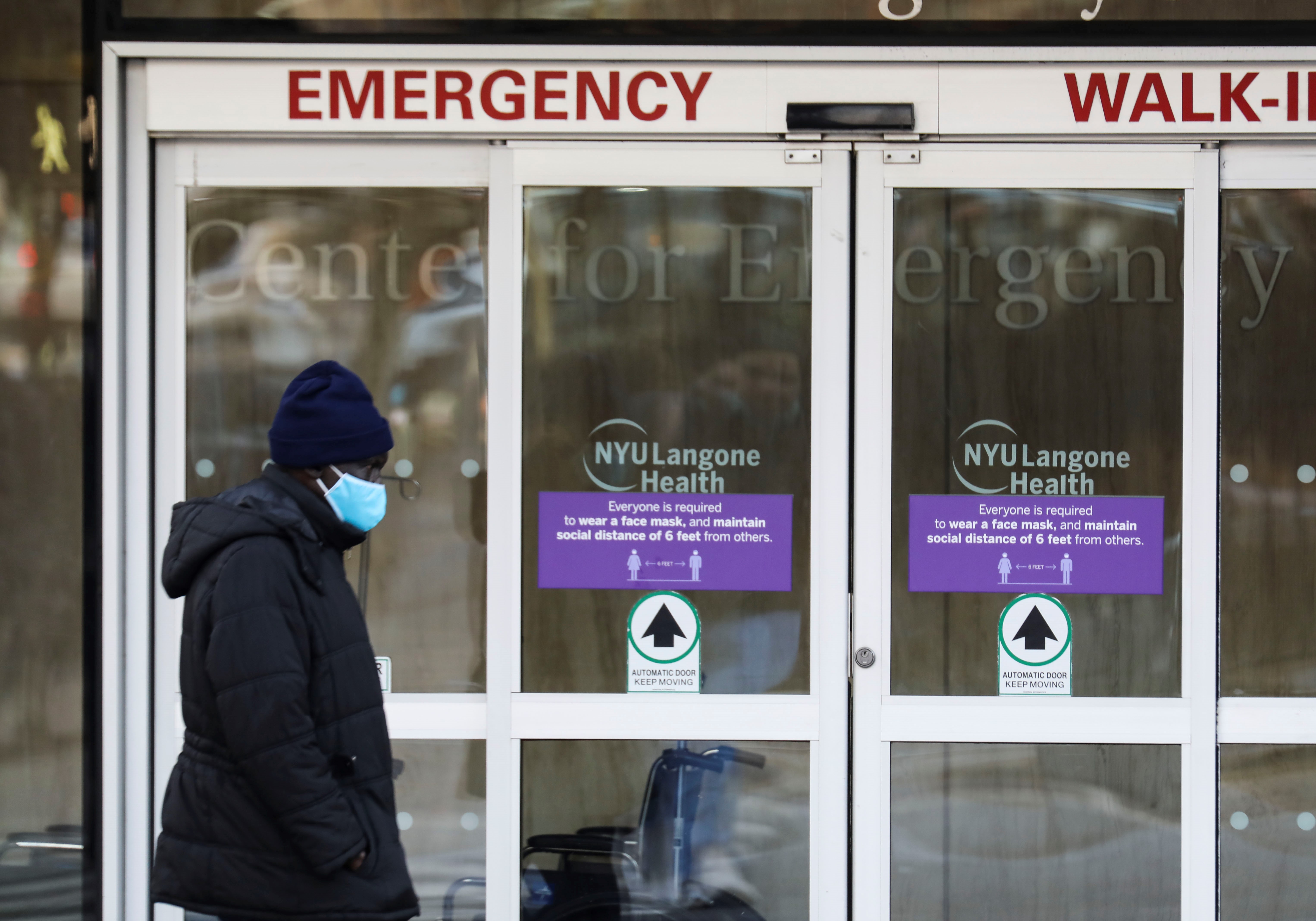 A person in a winter coast and face mask walking past the entrance of a hospital emergency department.