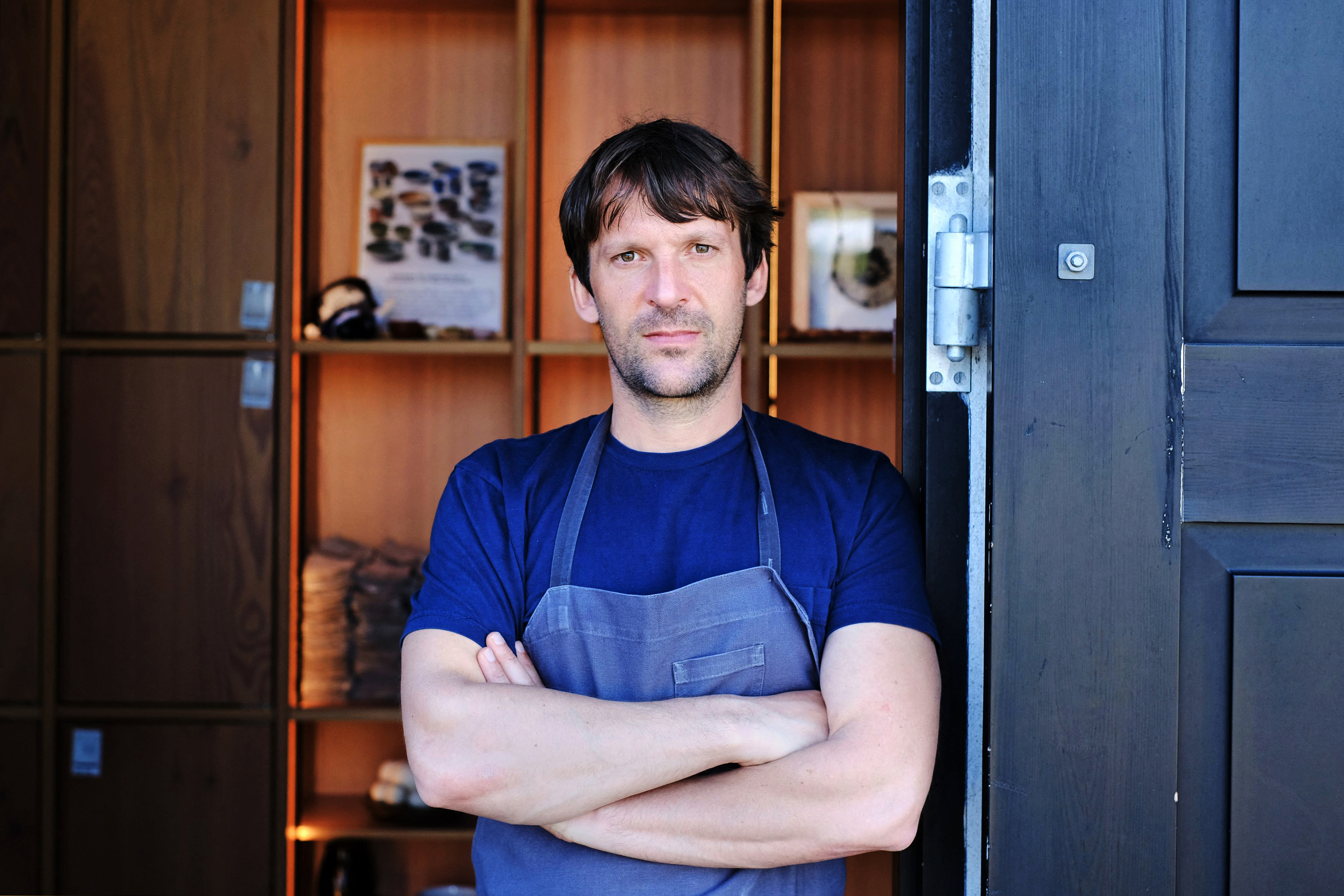 Rene Redzepi stands in a doorway, wearing an apron and crossing his arms.