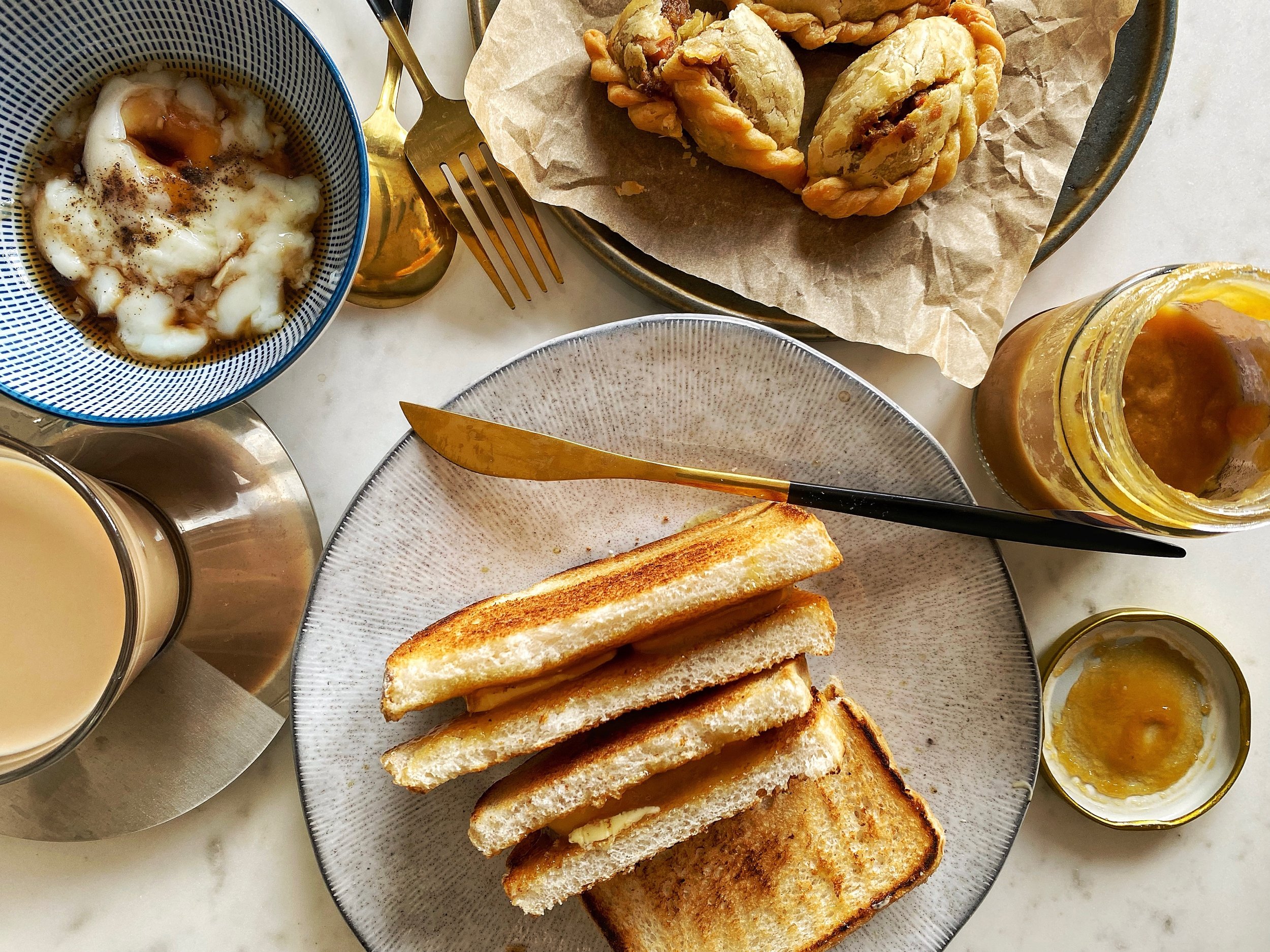 A birdseye view of a plate of kaya toast, a curry puff on greaseproof paper, and coddled eggs in a bowl with soy sauce.