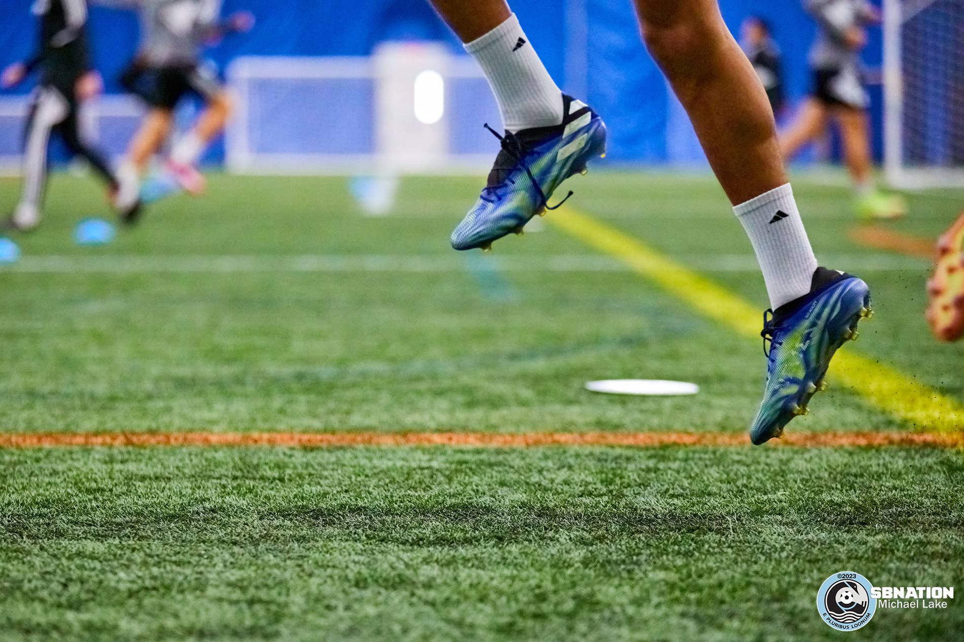A close-up of a pair of blue Adidas cleats are in focus as their owner skips in warmups, with other cleated feet out of focus in the background.