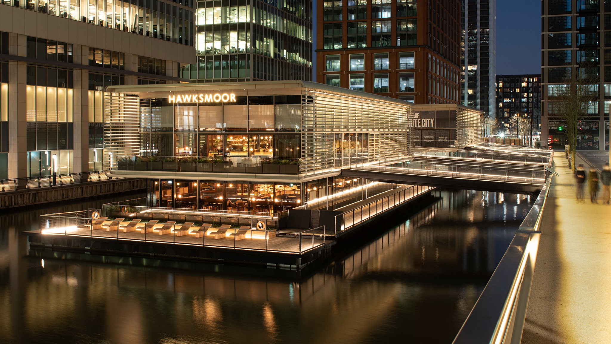 A floating restaurant and bar on three levels in Canary Wharf, with skyscrapers all around, at night.