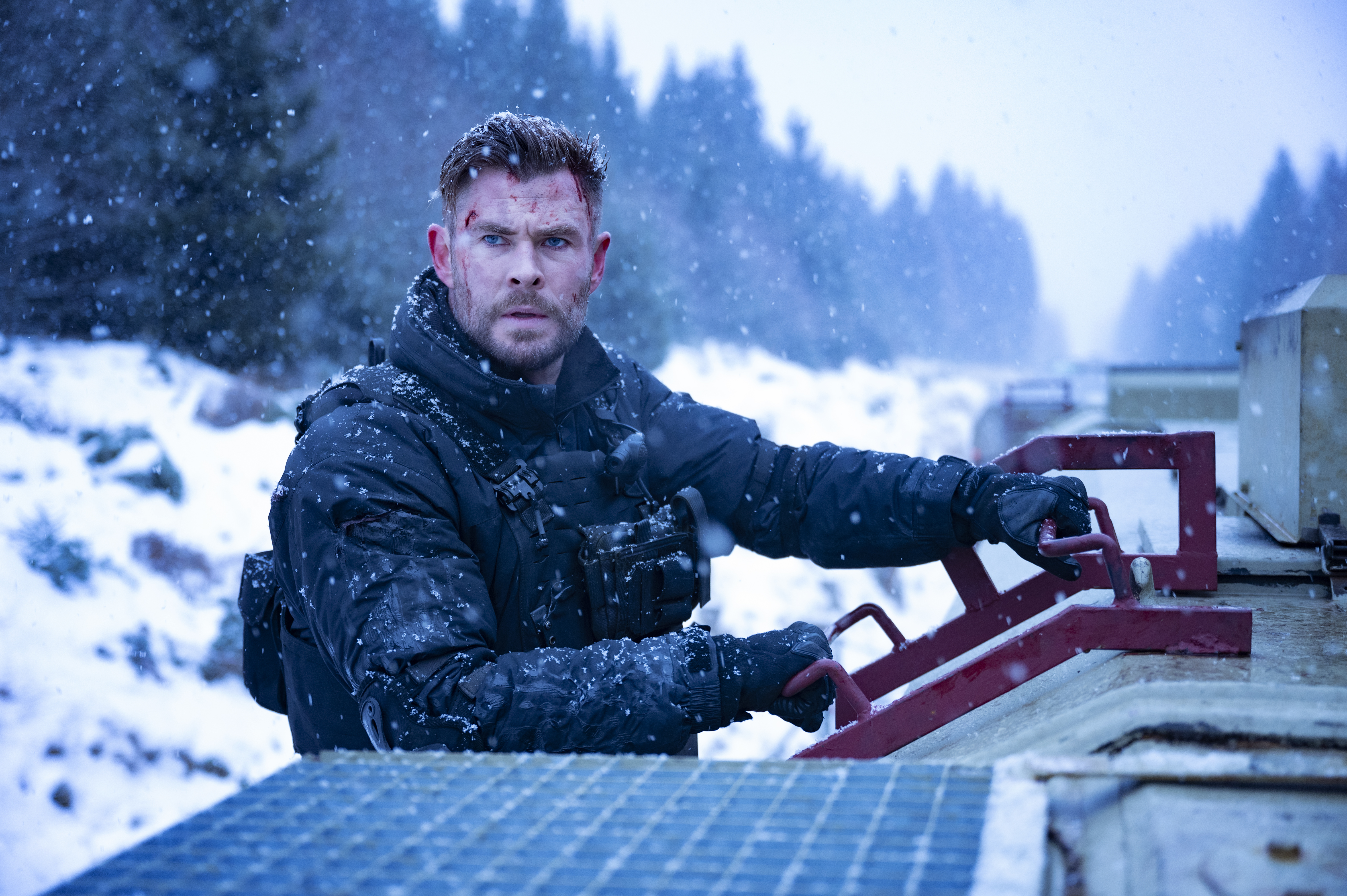 chris hemsworth in the snow. he stands next to a vehicle and looks determined