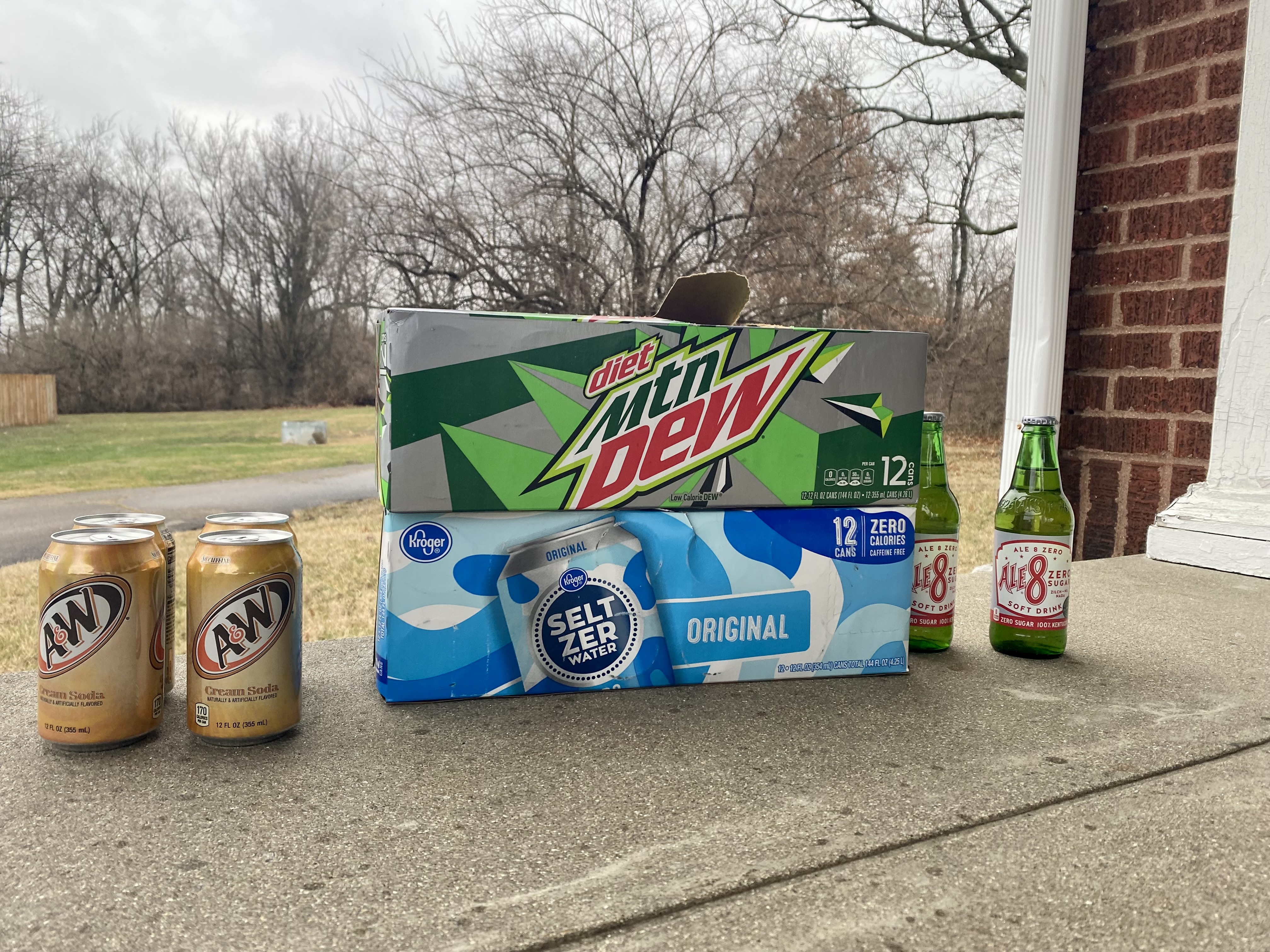 A few cans of A&amp;W root beer, boxes of Mountain Dew and seltzer, and two beers sit on a porch.