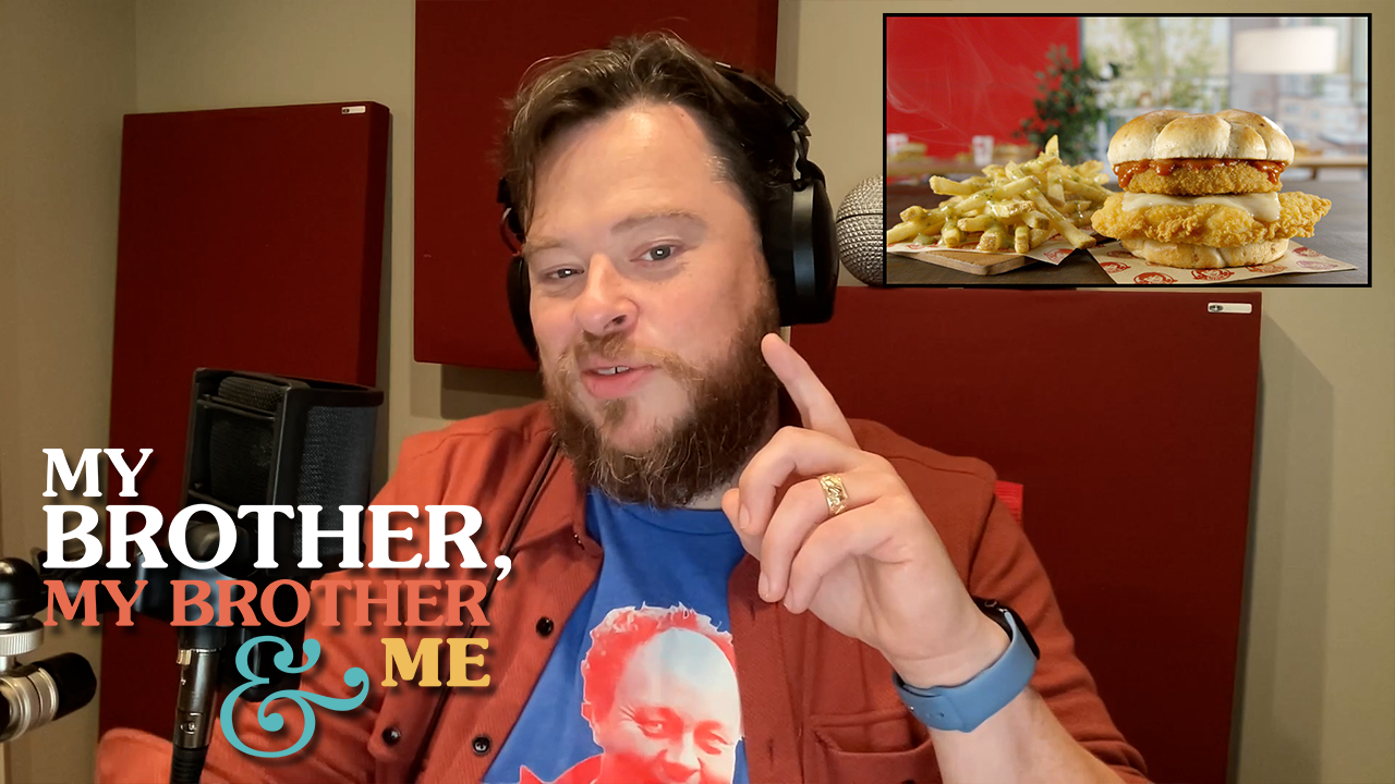 A photograph of Justin sitting at his desk in front of a microphone, wearing headphones and an orange button own shirt open over a blue tee. His left hand is raised. In the upper right corner is a superimposed photo of a fried chicken sandwich with an additional fried patty on top of it, and french fries on the side. In the lower left corner is the logo for My Brother, My Brother and Me.