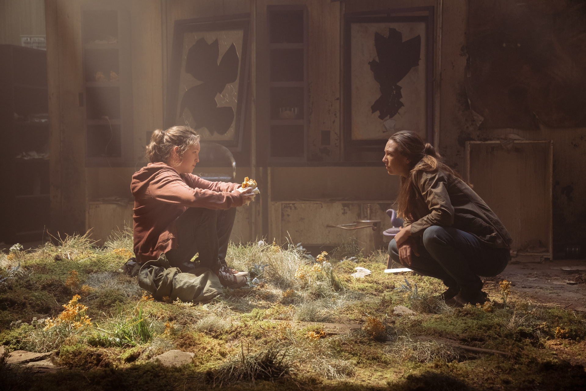 Ellie and Tess crouch facing each other in the grass that has overtaken a decrepit building as light shines in from above in the HBO series The Last of Us.