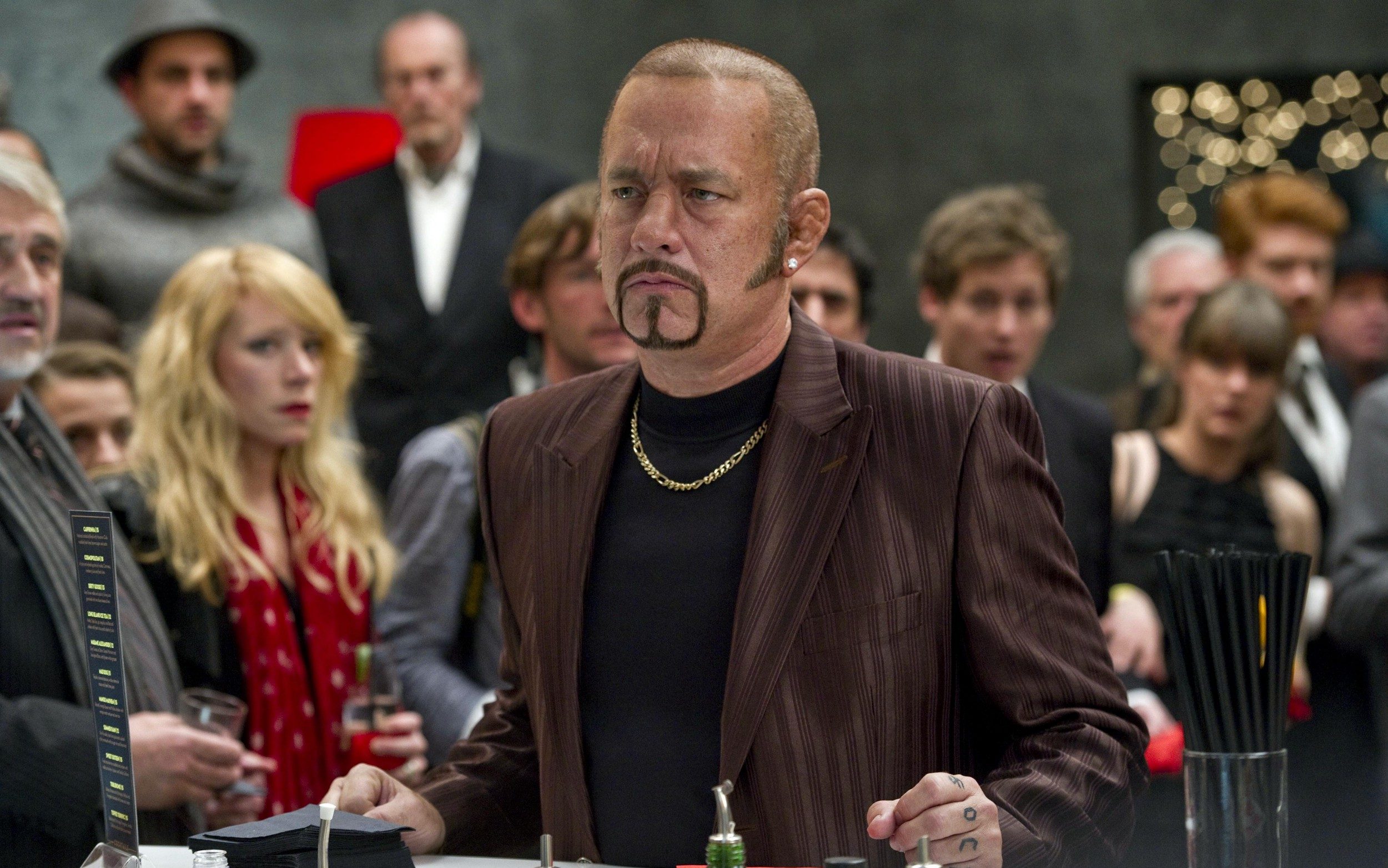 Tom Hanks with a sharp beard, gold necklace, earings, and a red blazer stands in front of a crowd who all seem intimidated by him in Cloud Atlas