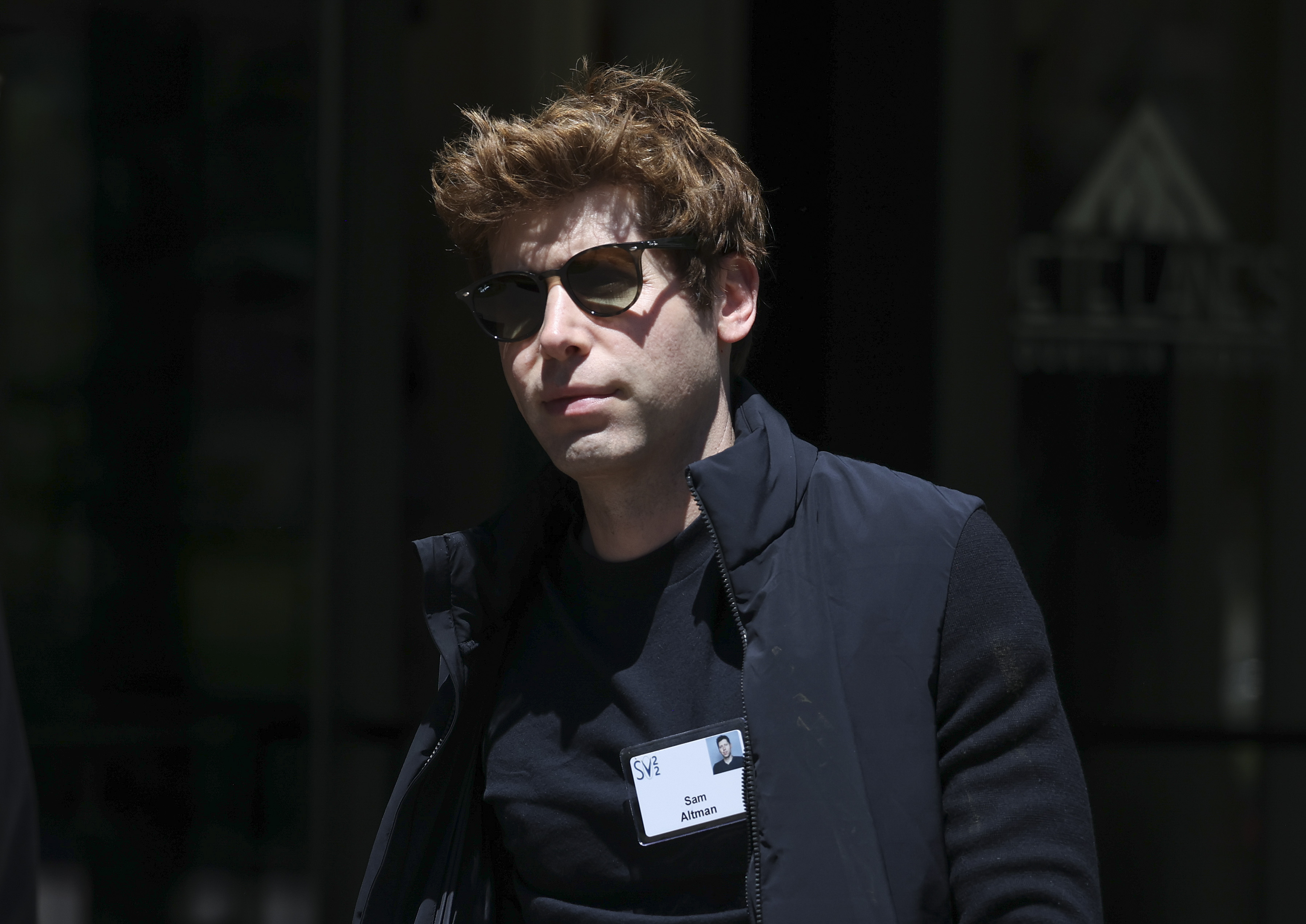 A close up of OpenAI CEO Sam Altman wearing a black t-shirt and black sunglasses.