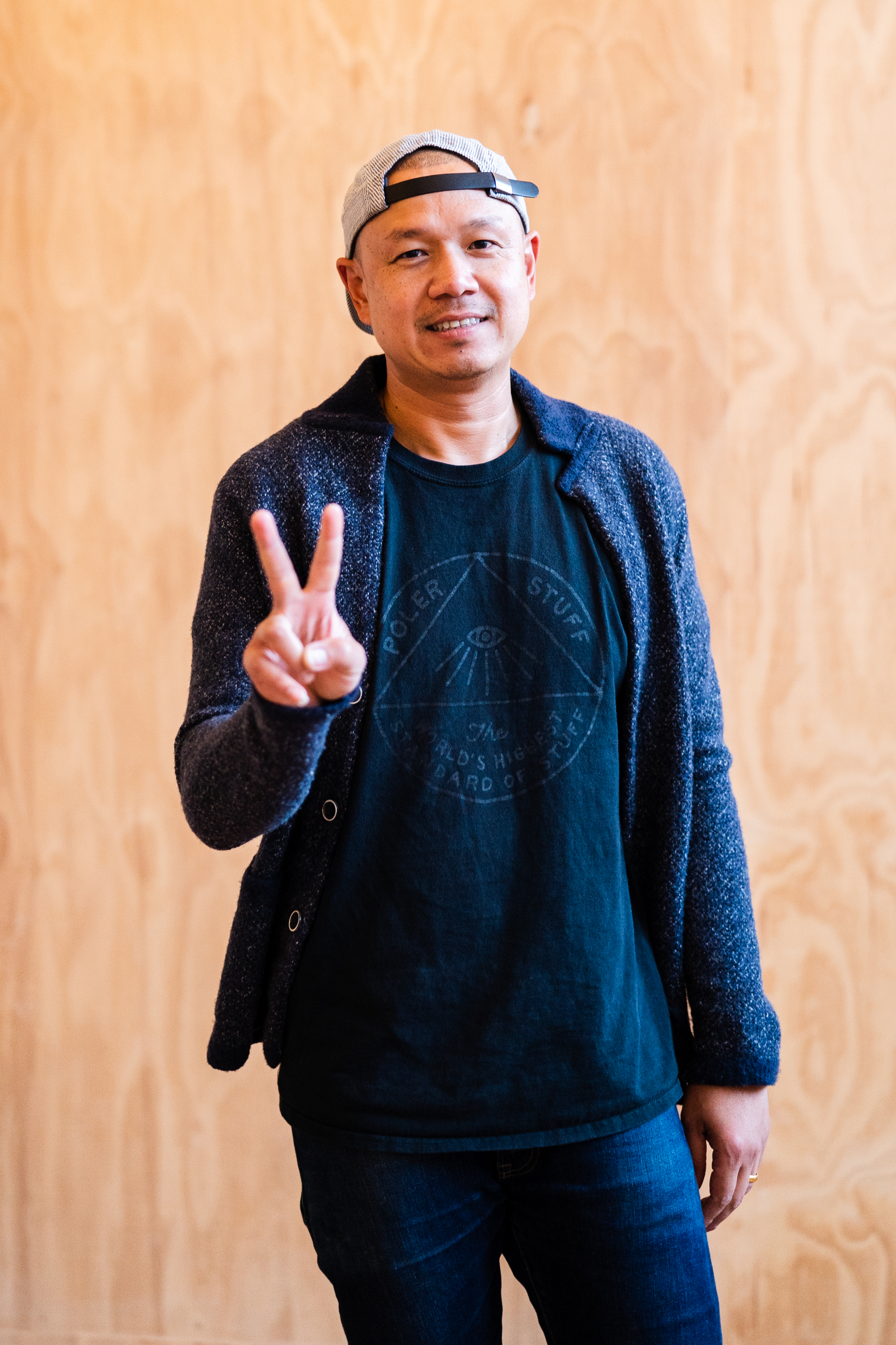 A 40-year-old Earl Ninsom stands in front of a wooden wall, holding up a peace sign. He’s wearing a loose jacket, a t-shirt, and a backwards snapback cap, with a smile.