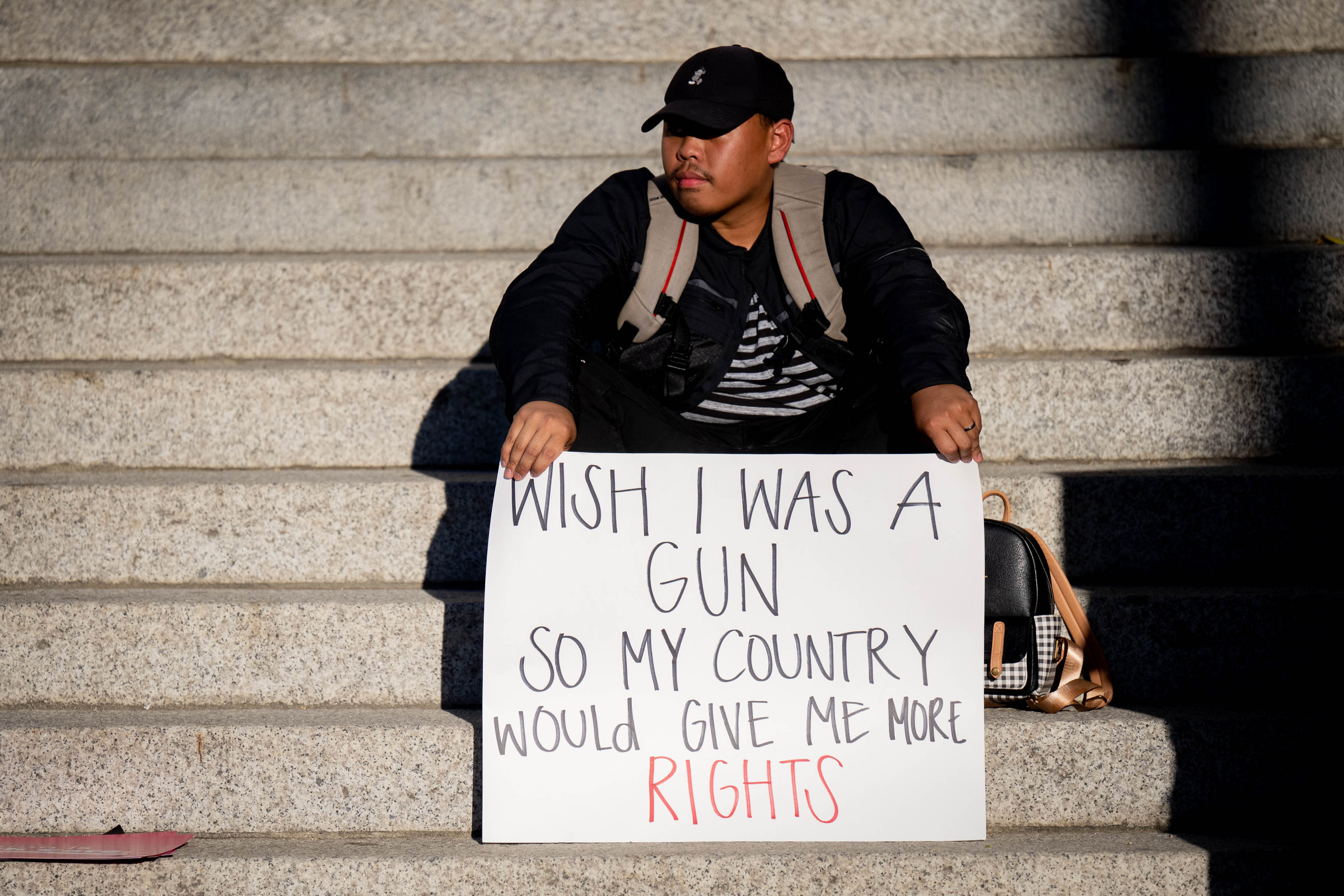 A man in a baseball cap sits on a flight of stairs with a sign propped on his knees reading “Wish I was a gun so my country would give me more rights.” 