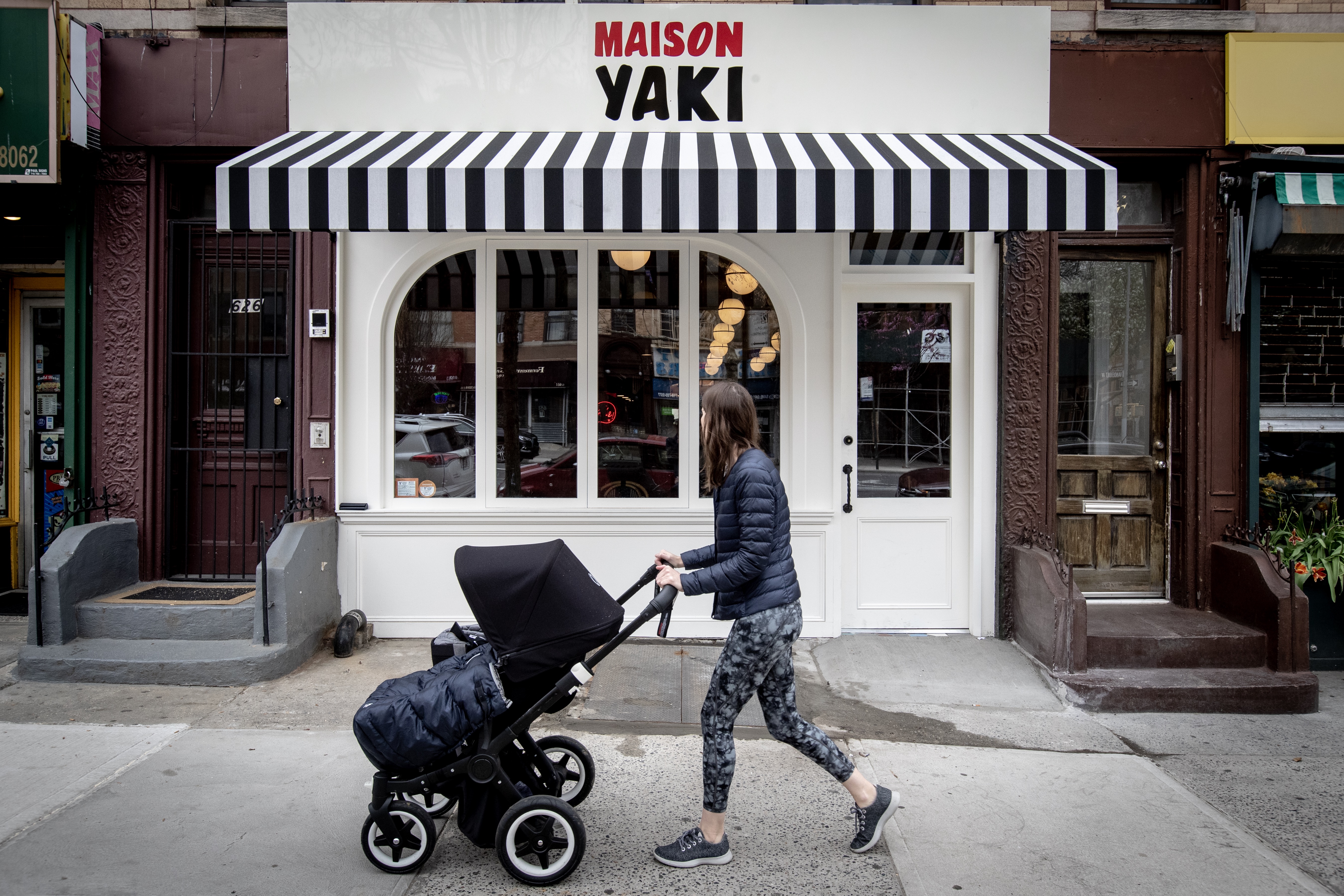 A person with a stroller passes in front of a white restaurant with a striped awning.