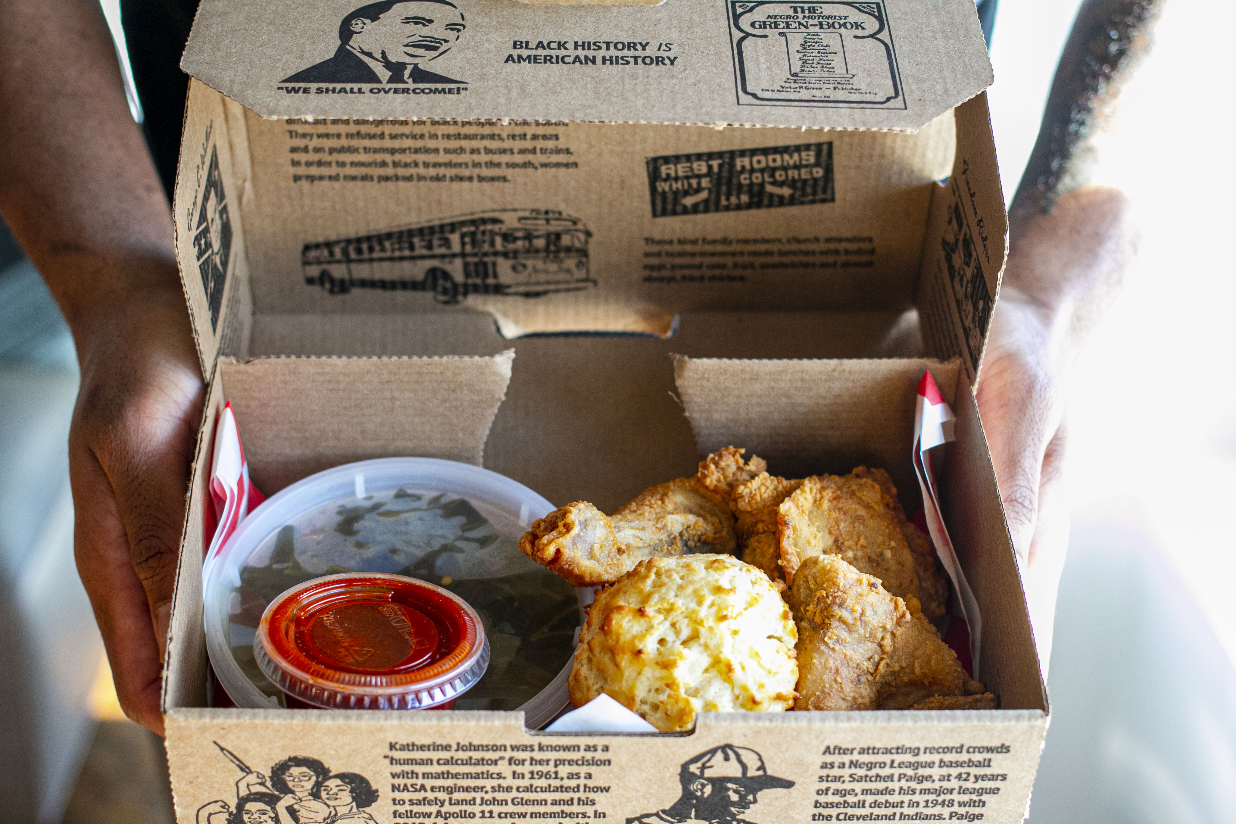 A cardboard box filled with chicken, a biscuit, order side dishes, held by two hands.