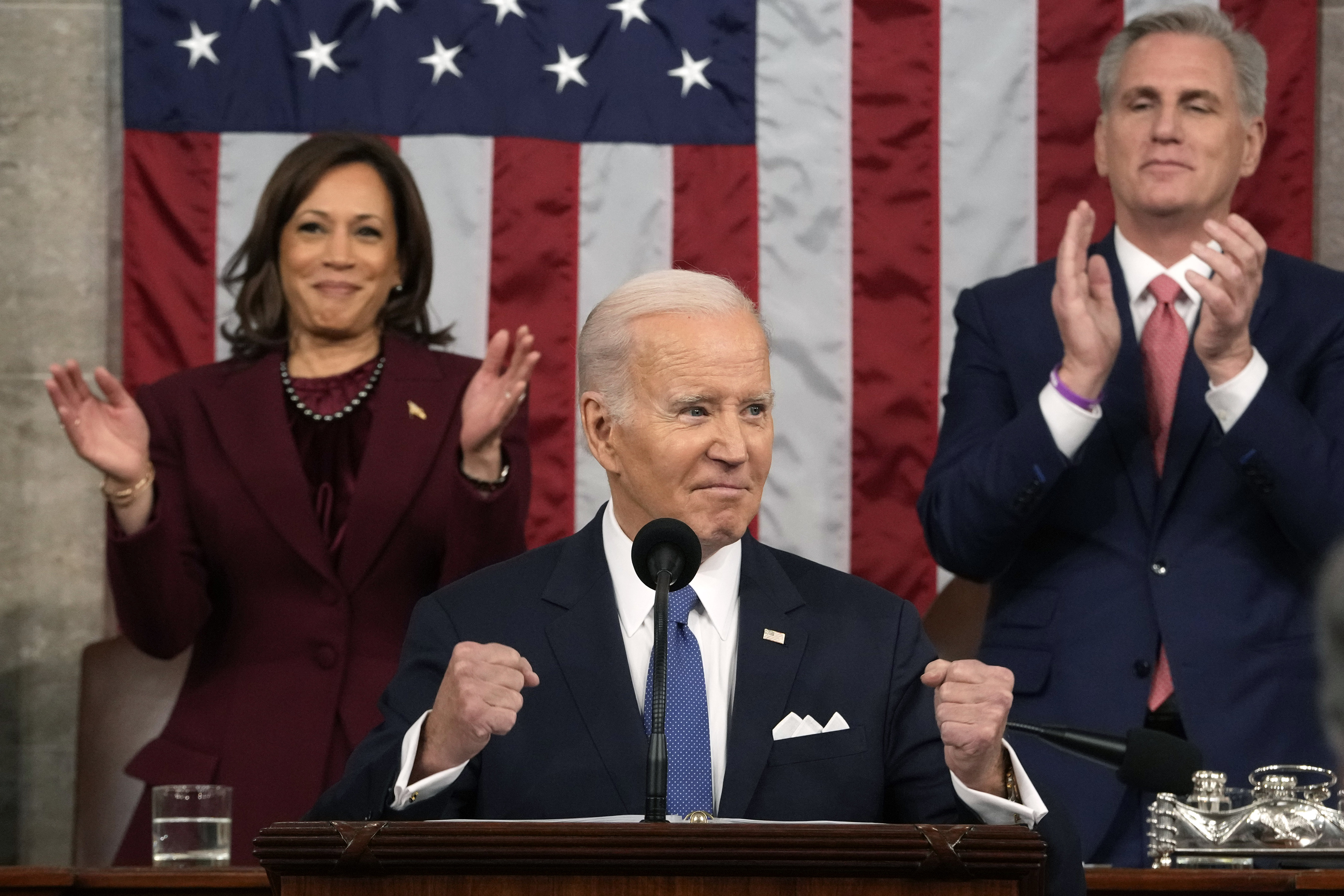Biden, in a navy suit, white shirt, and blue tie, pumps his fists for emphasis as a ruby suited Vice President Harris and a navy suited House Speaker Kevin McCarthy stand behind him and applaud. 