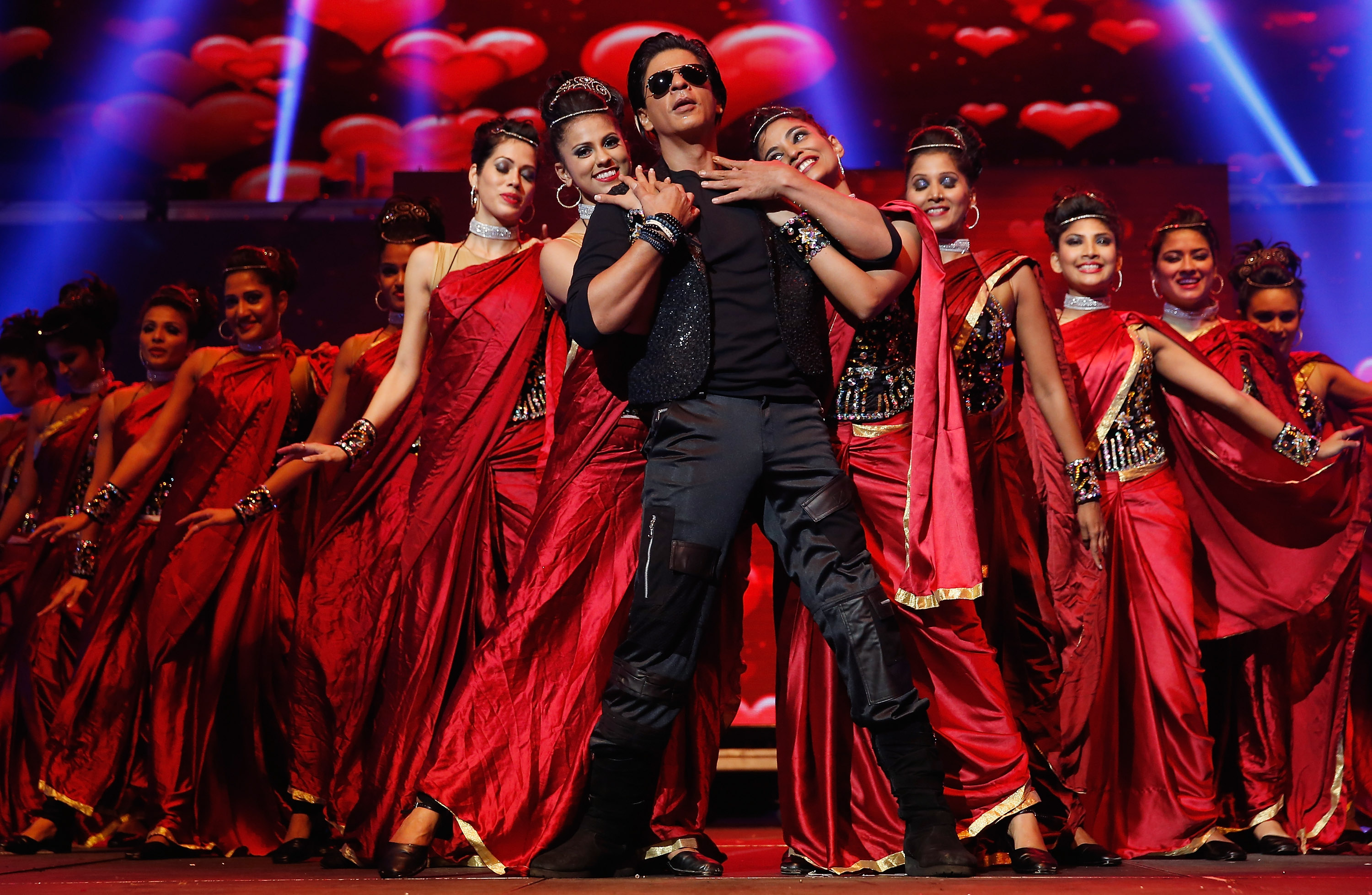 Bollywood actor Shah Rukh Khan onstage wearing dark clothes and sunglasses, flanked by women dancers in red saris. 