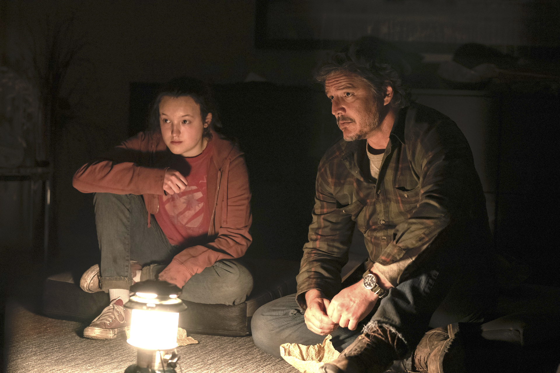 Ellie (Bella Ramsey) and Joel (Pedro Pascal) sit on the floor together, lit by a lantern in front of them in The Last of Us.