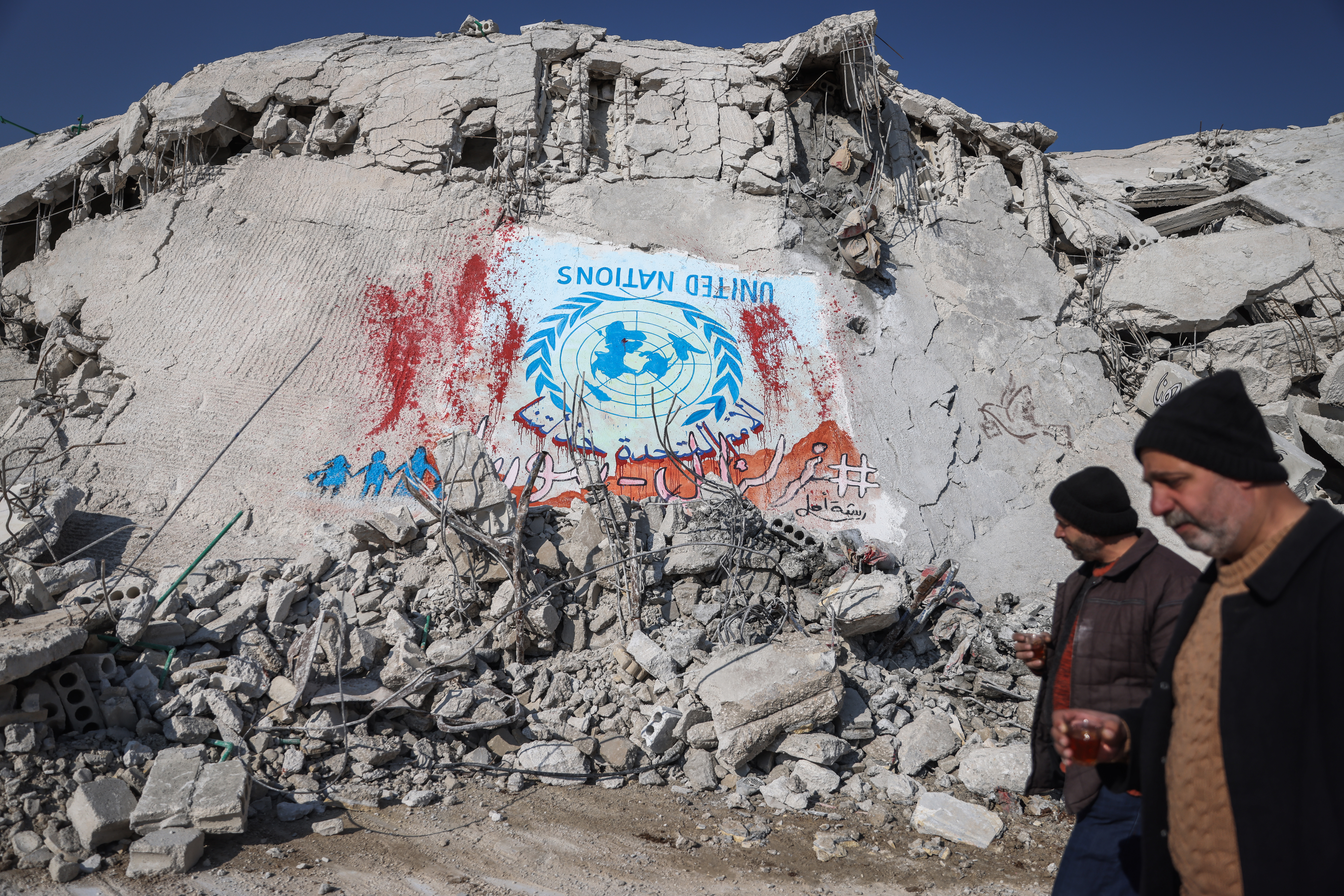 People in Sarmada, Syria, walk past rubble. A blue and white United Nations flag is painted upside down on a destroyed building.