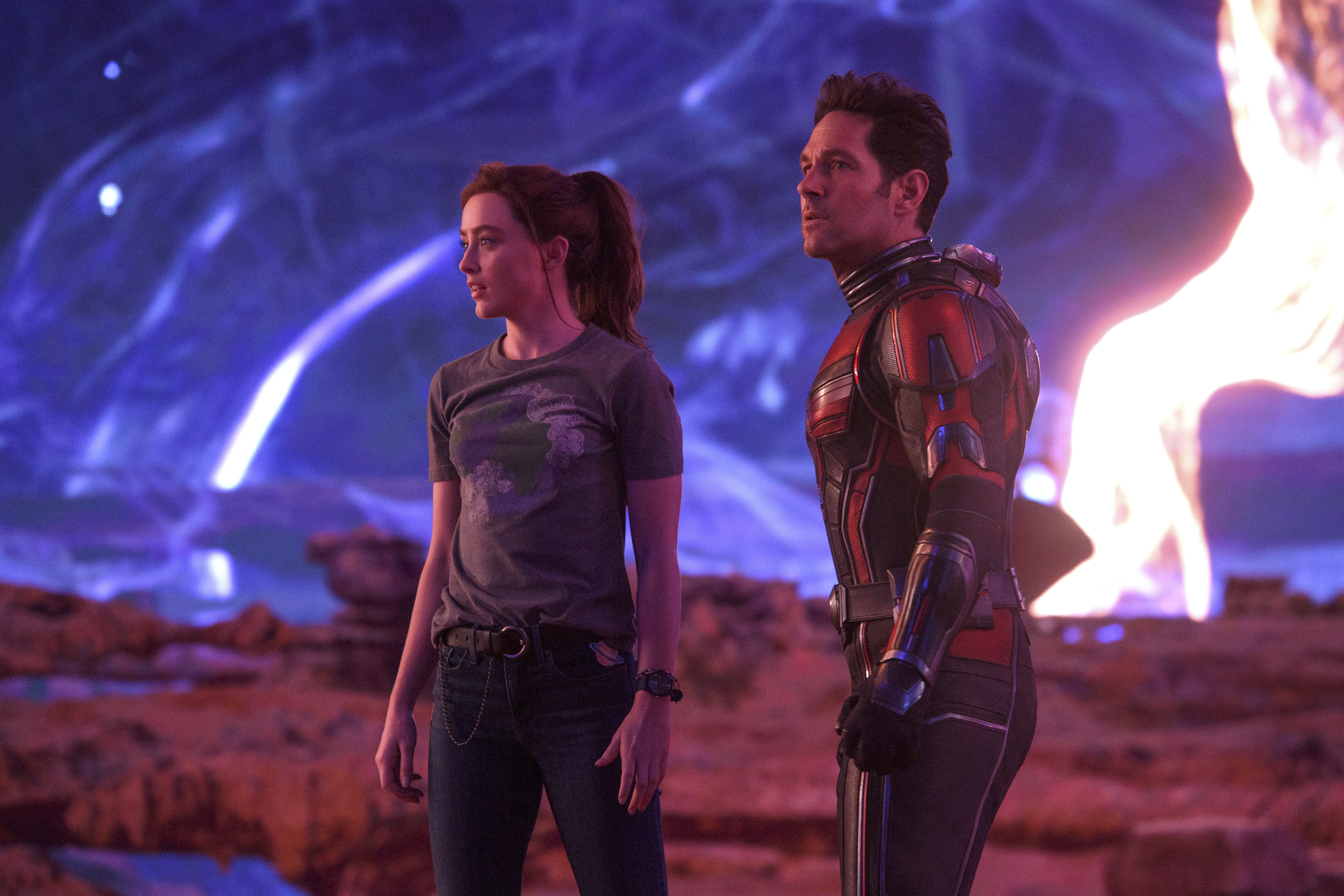 (L-R): Kathryn Newton as Cassandra “Cassie” Lang and Paul Rudd as Scott Lang/Ant-Man in Ant-Man and the Wasp: Quantumania. Cassie is in jeans and a t-shirt, while Scott is wearing his Ant-Man costume with the helmet off. They stand stunned on an alien vista with swirling skies.