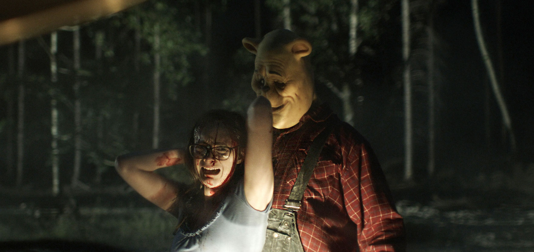 A grown-up Winnie the Pooh (Craig David Dowsett, in a vinyl mask) holds a wailing, bloodied victim by the hair in the light of a truck’s headlights in Winnie the Pooh: Blood and Honey
