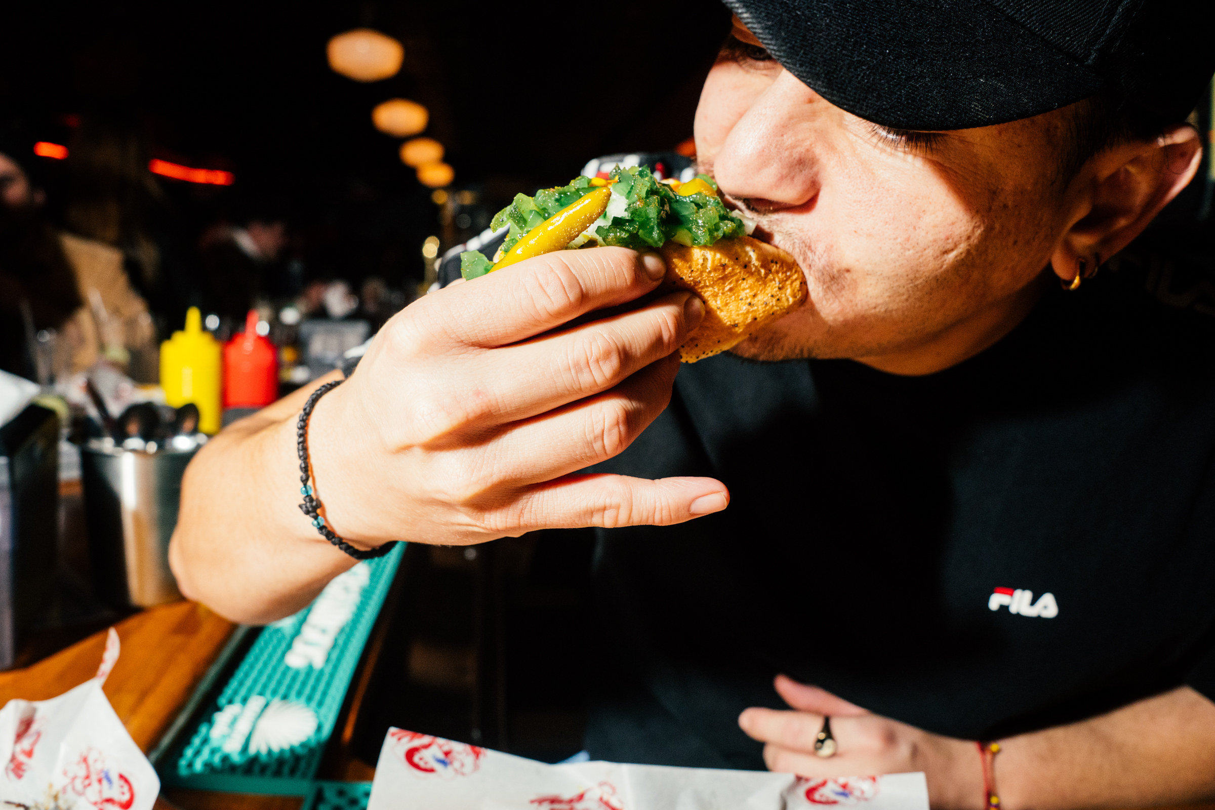An employee at a bar takes a bite of a Chicago-style hot dog with relish and sport peppers.