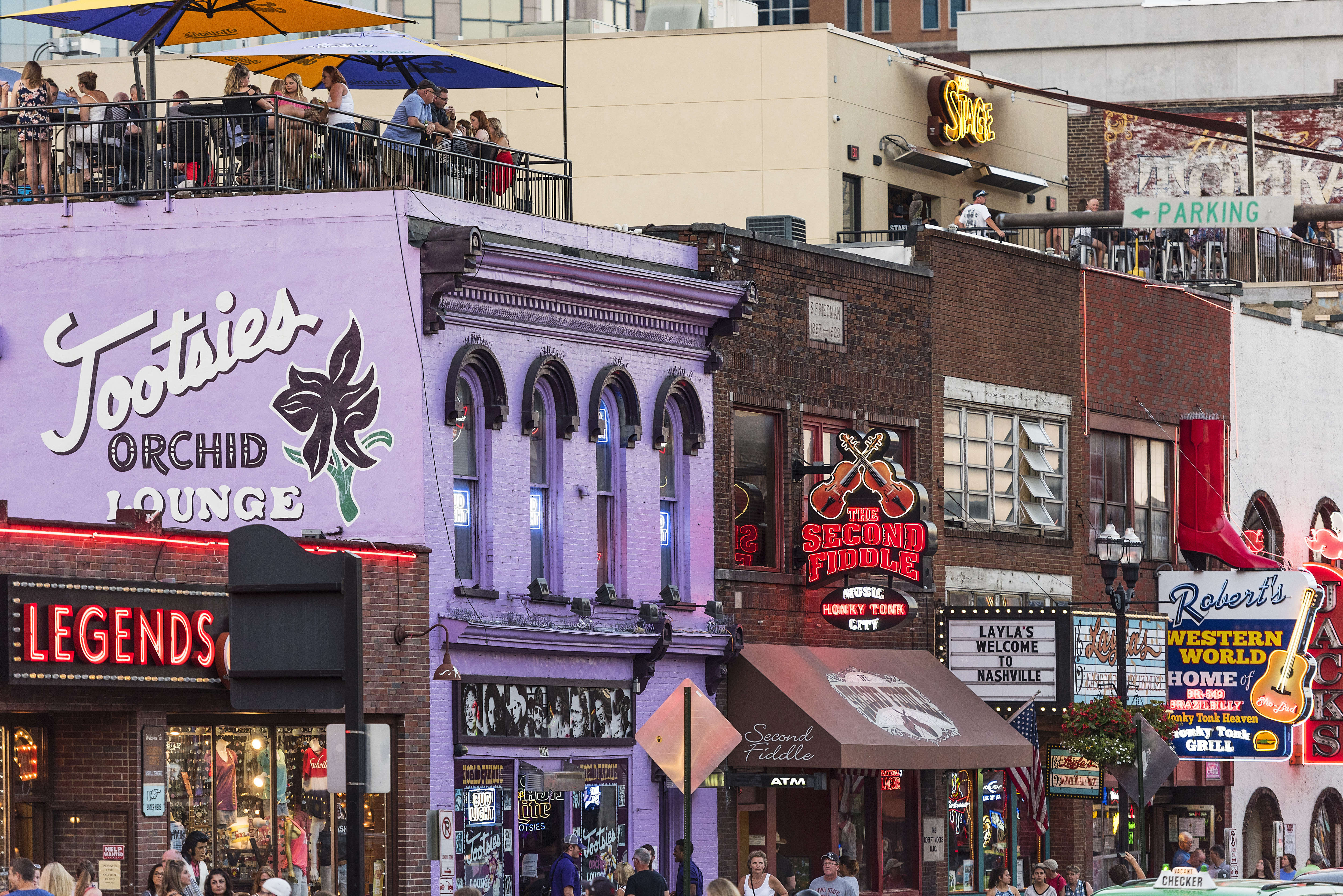 The exterior of several buildings on Broadway in Nashville, including a purple one with Tootsies Orchid Lounge painted on the side, one with a neon Legends sign out front, and one with a neon Second Fiddle sign out front.