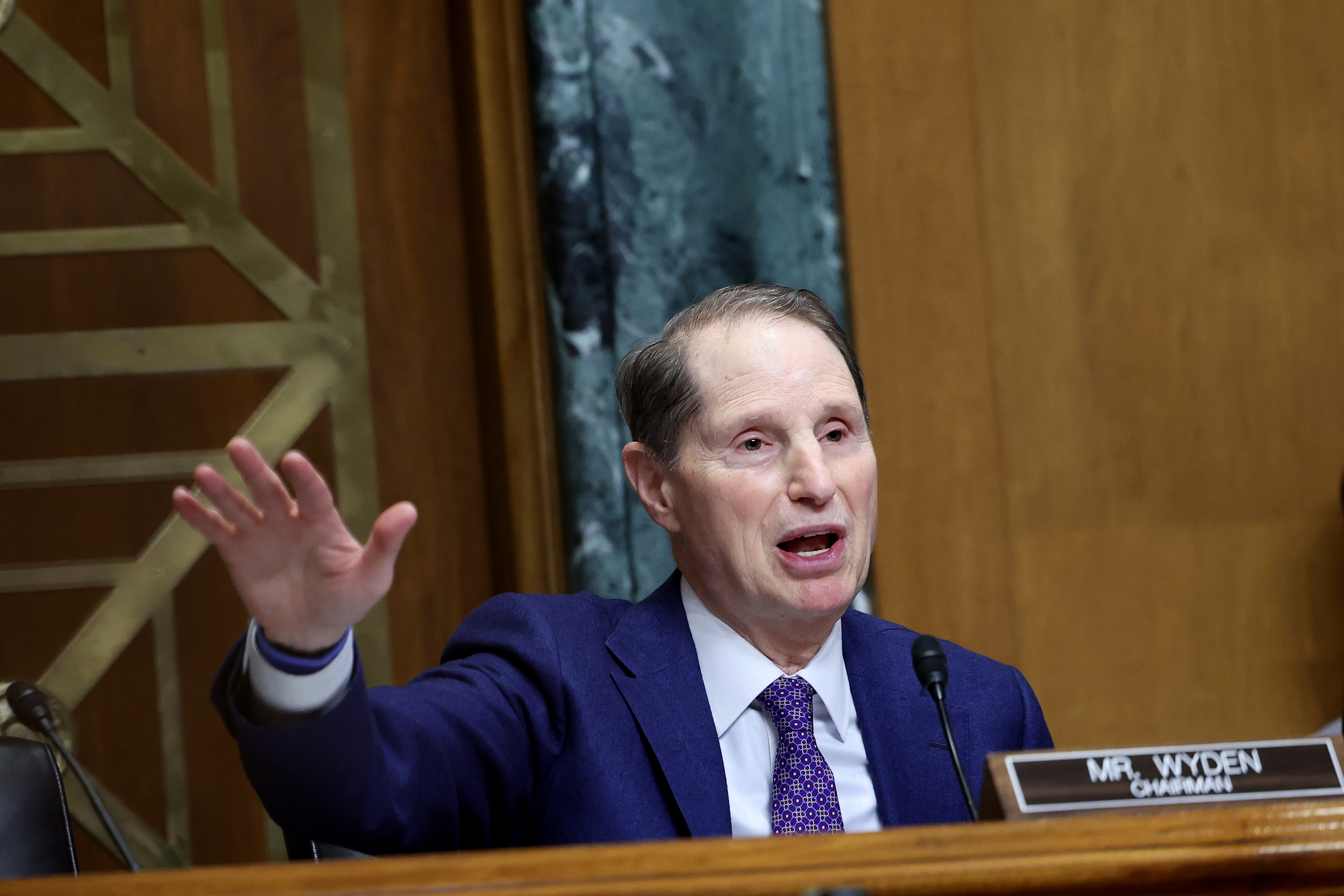Senator Ron Wyden, speaking from behind his nameplate and gesturing out with his right hand.