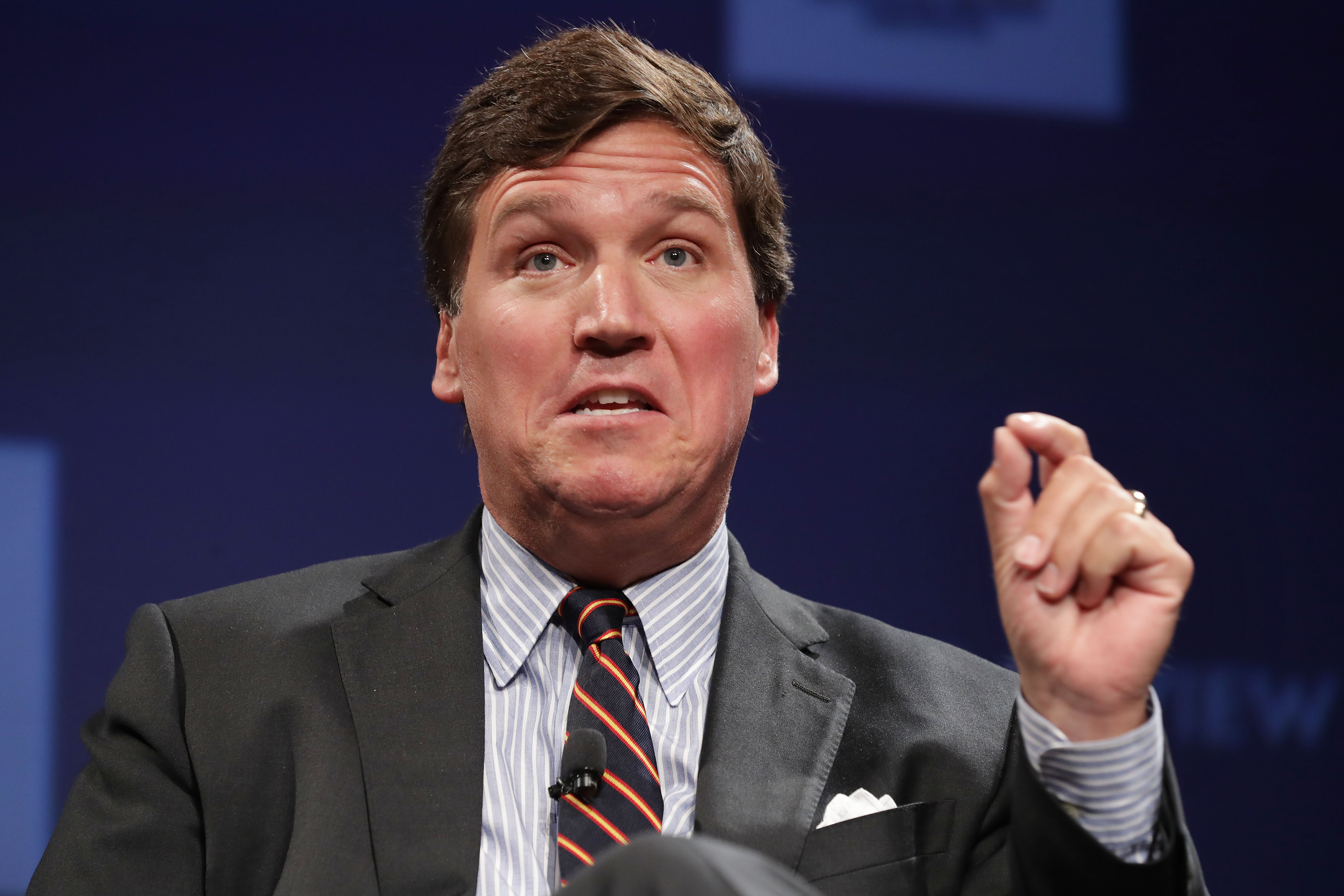 Tucker Carlson speaks, gesturing with one hand, to an unseen audience. He’s wearing a dark gray suit, a white-and-gray-striped shirt, and a navy striped tie, and sits in front of a dark blue background.