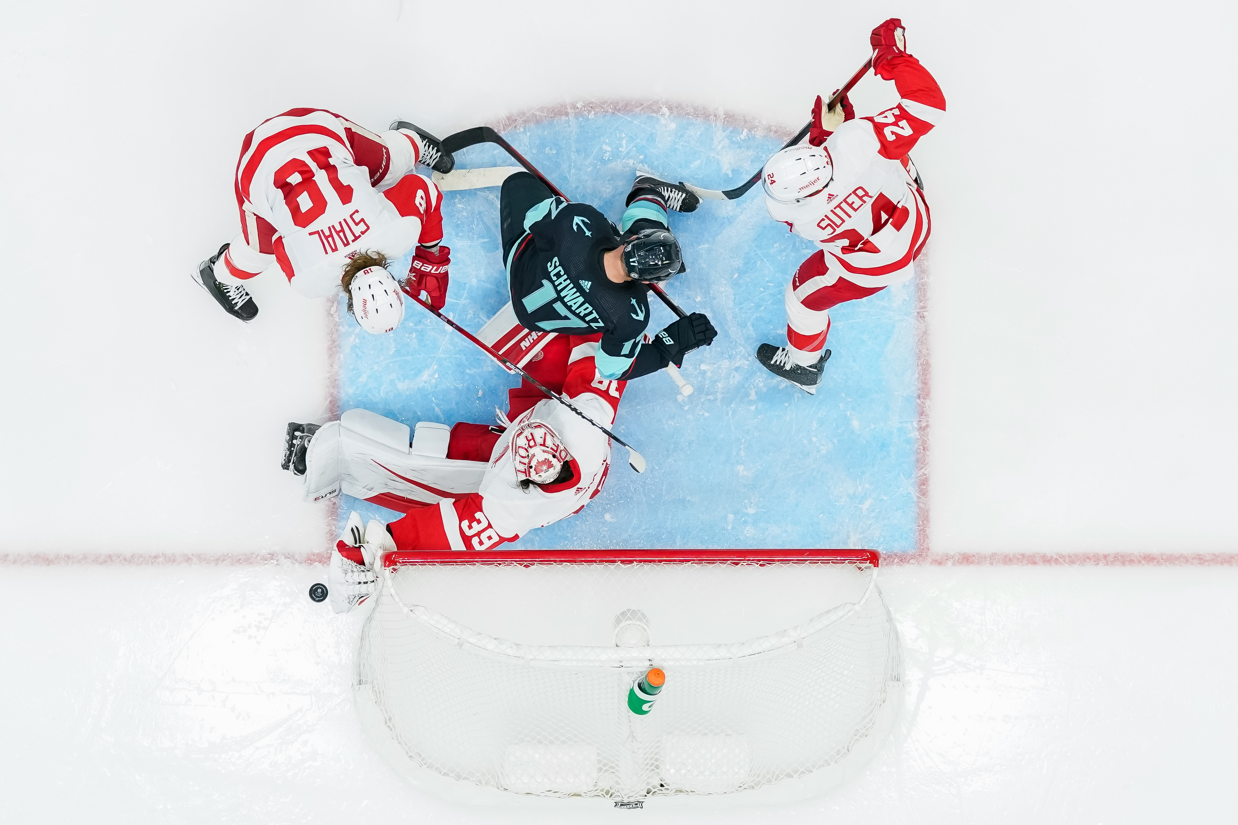 An overhead shot above the Detroit net. Jaden Scwhartz is in the middle of the bluepaint trying to score surrounded by two Red Wings on either side, but the Detroit netminder is reaching out to make the save. Scwhartz is in the navy blue home jersey and the Detroit players are in their white away jerseys.