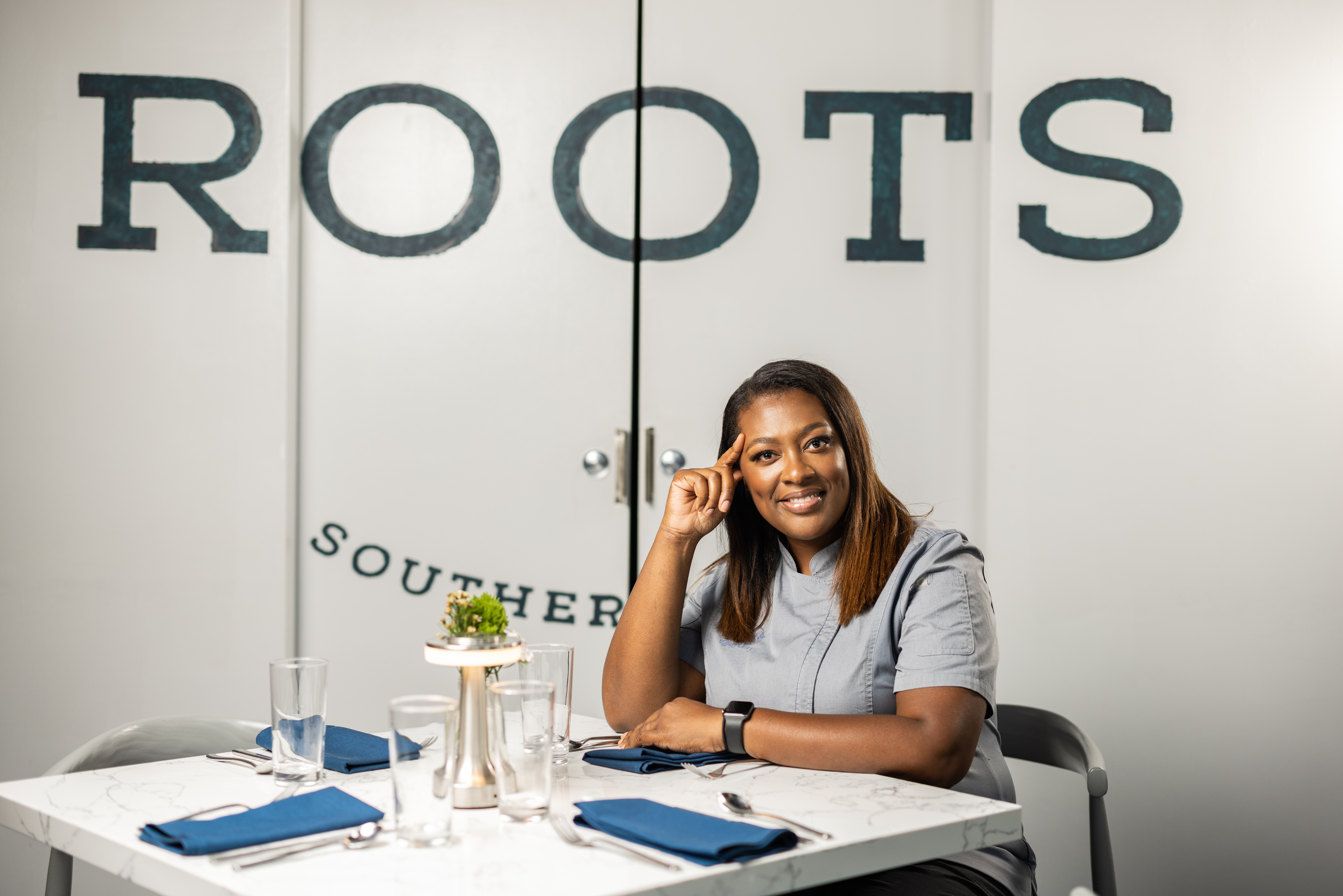 A chef sits at the table. Behind her on a white wall, the words “Roots Southern” are printed in navy.