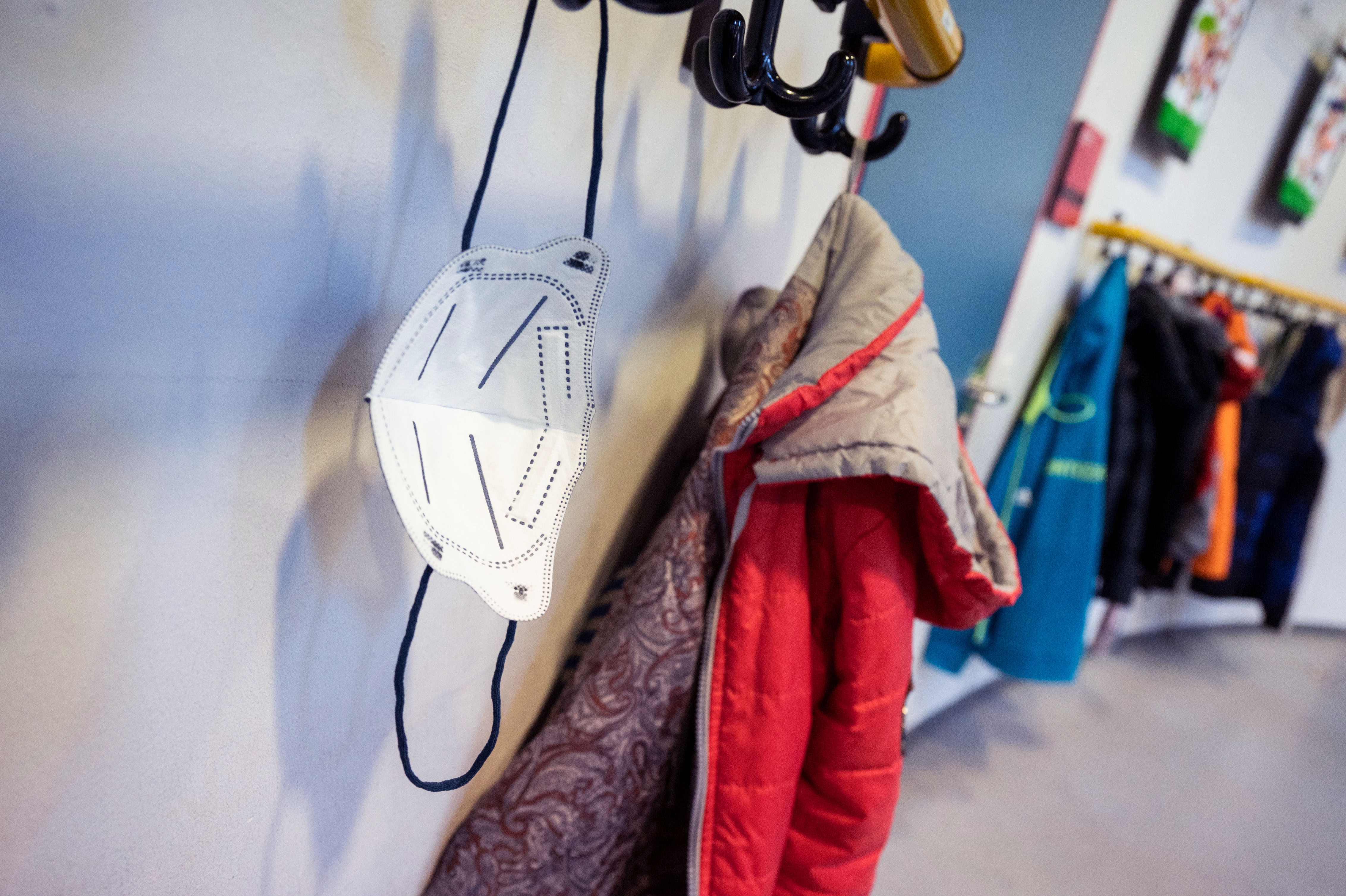 In a school hallway, a white mask hangs on a row of hooks next to a red child’s coat, with more coats in the background.