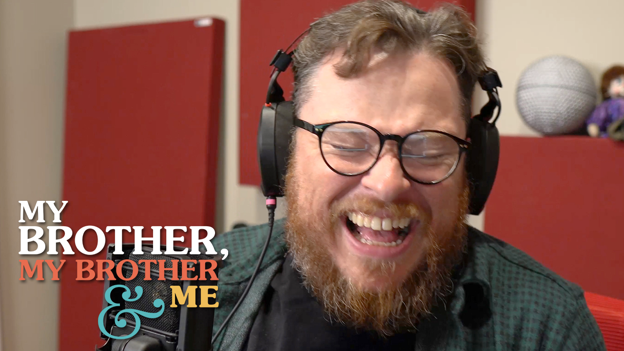 A screencap of Justin sitting at his desk, laughing. He is seated in front of a microphone, wearing headphones and glasses. The My Brother, My Brother and Me logo is superimposed in the lower left corner.