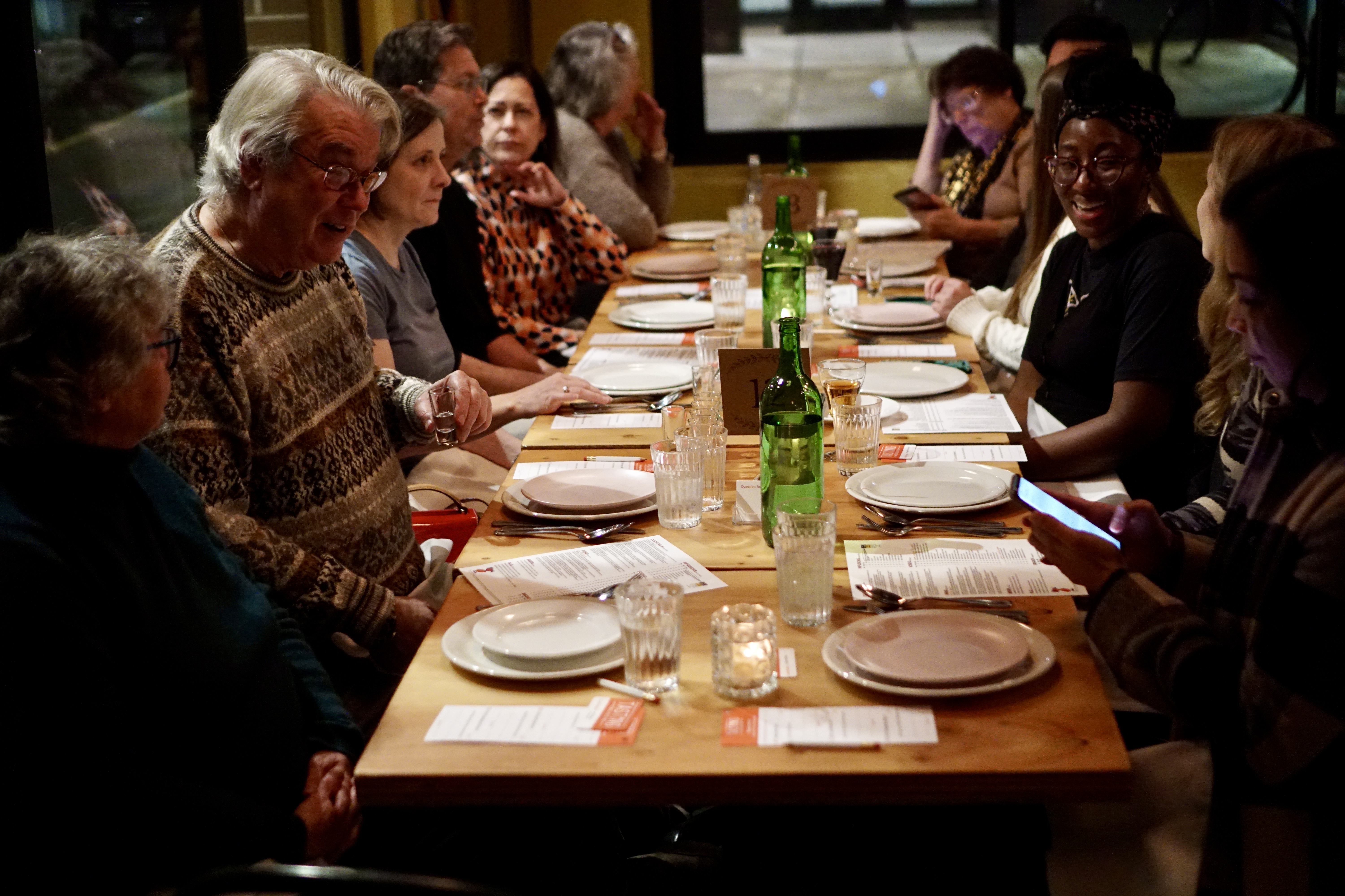Diners sit at a long communal dinner table and interact over the table, which is set with plates and menus.