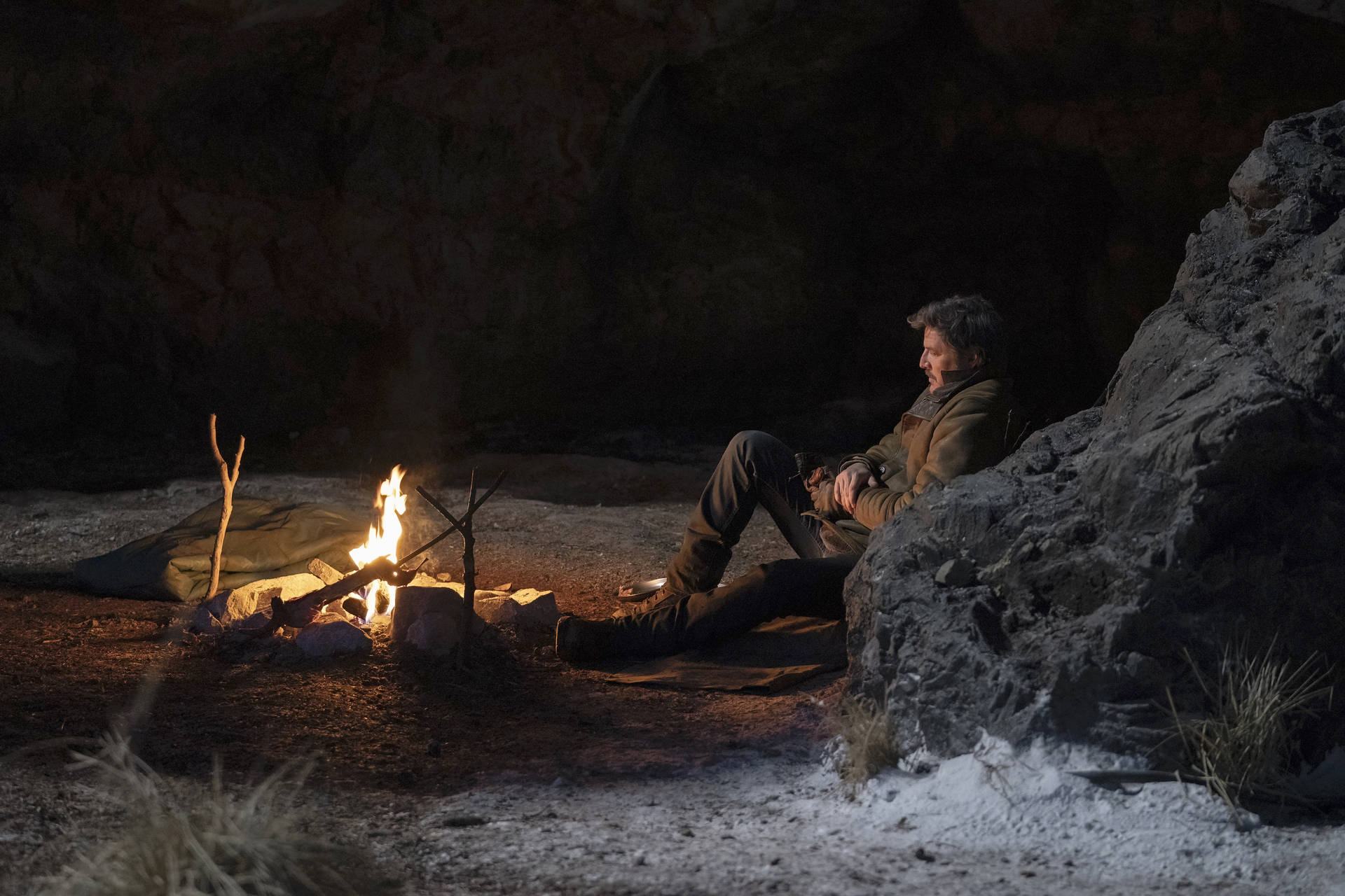 Joel (Pedro Pascal) sitting and leaning against a rock wall and looking at a fire
