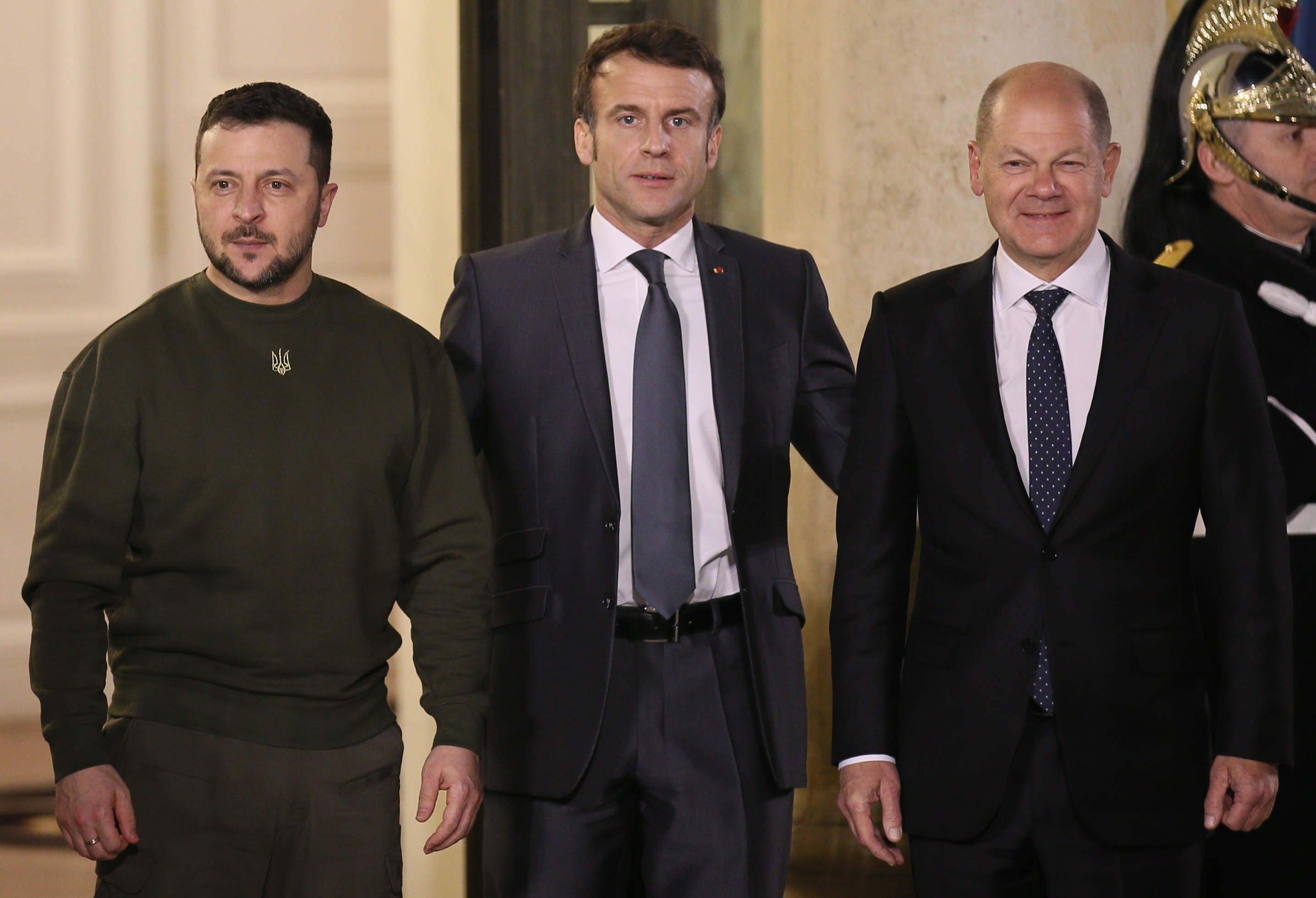 Left: President Zelenskyy wearing his signature army green sweatshirt and pants. Center: President Macron, wearing a black suit. Right: German Chancellor Scholz wearing a black suit and smiling.