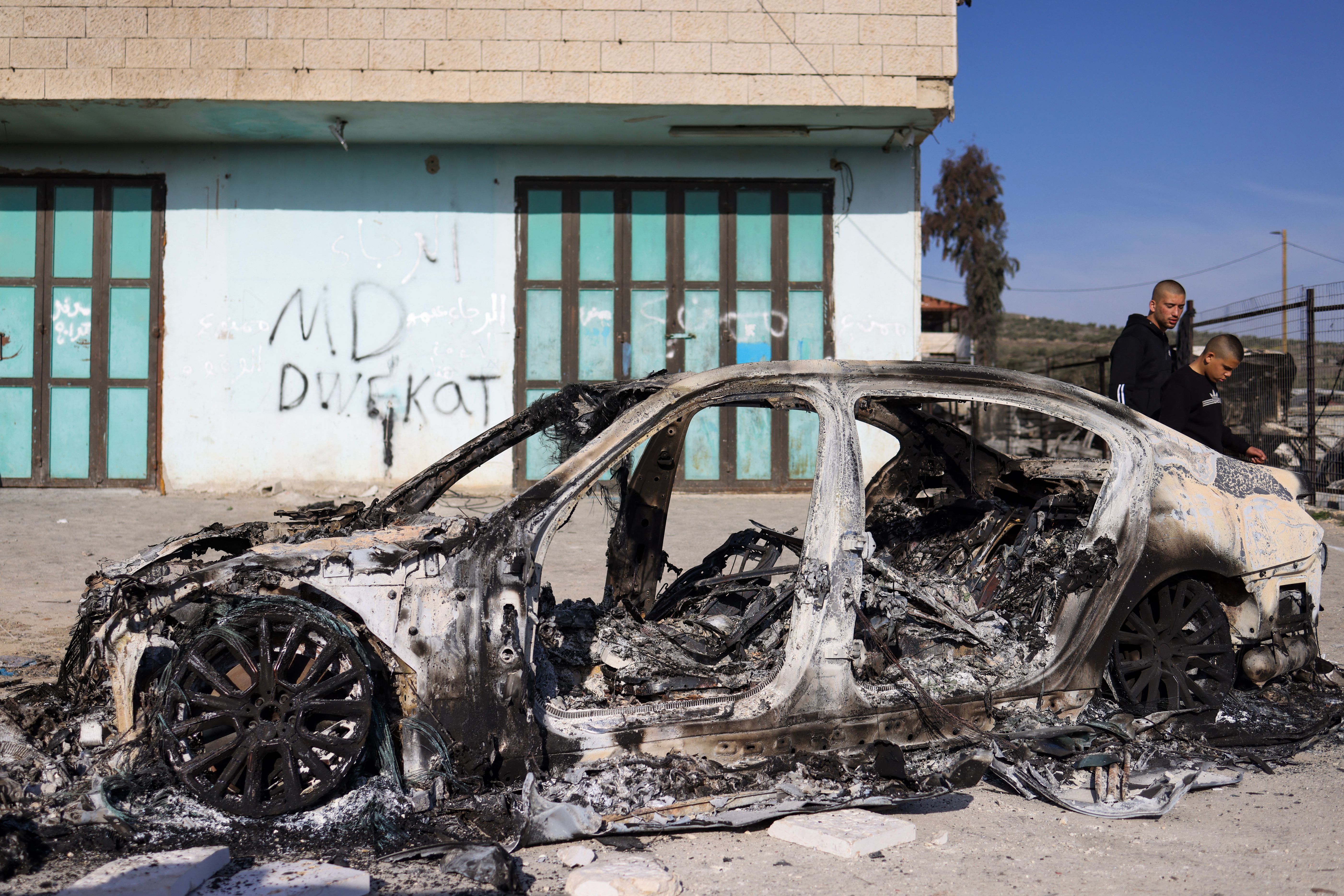A burned-out four-door car beside a graffitied building.