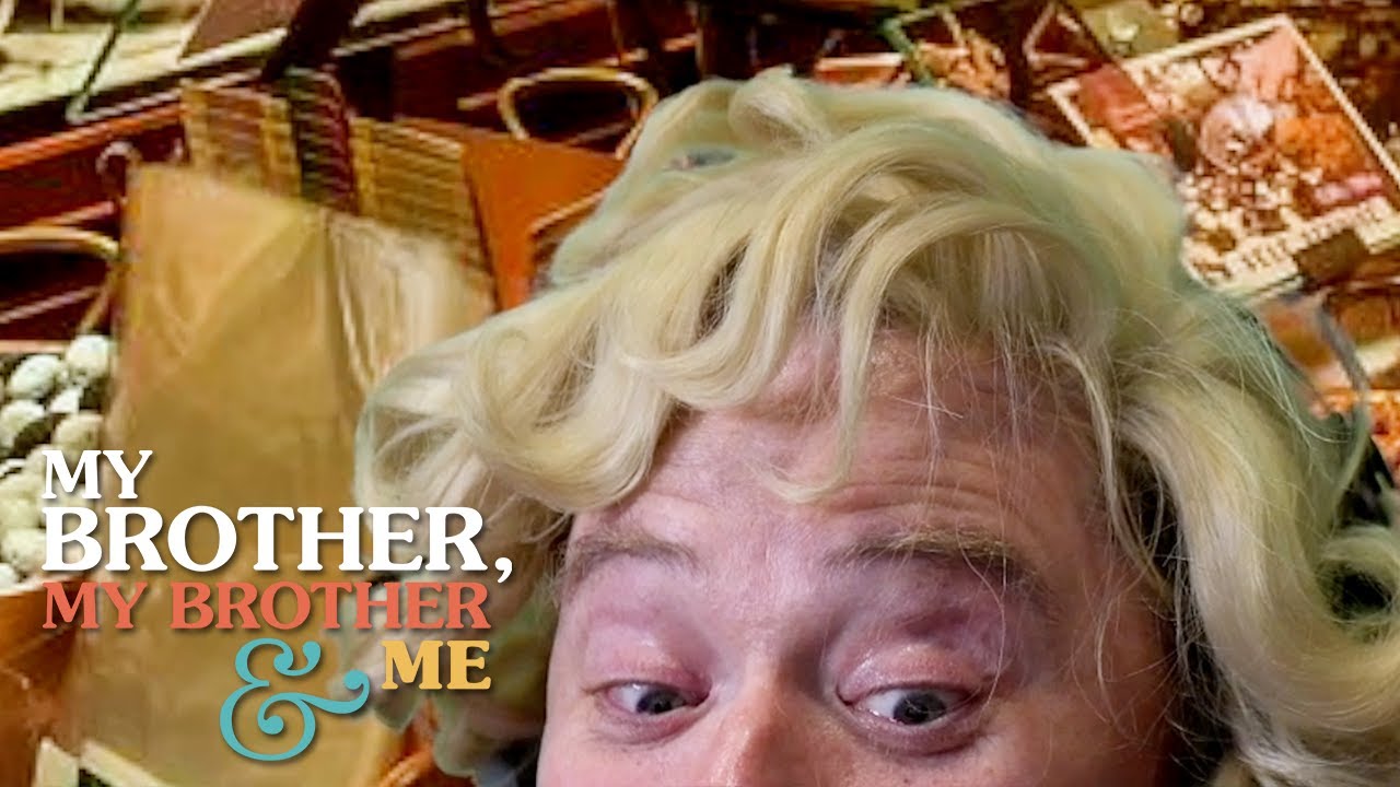 A screenshot of Justin McElroy peeking up from the bottom of a video frame. He is wearing a blonde curly wig, and his background is a photo of a shop with shelves full of candy and nuts. In the bottom left corner is the My Brother, My Brother and Me logo.