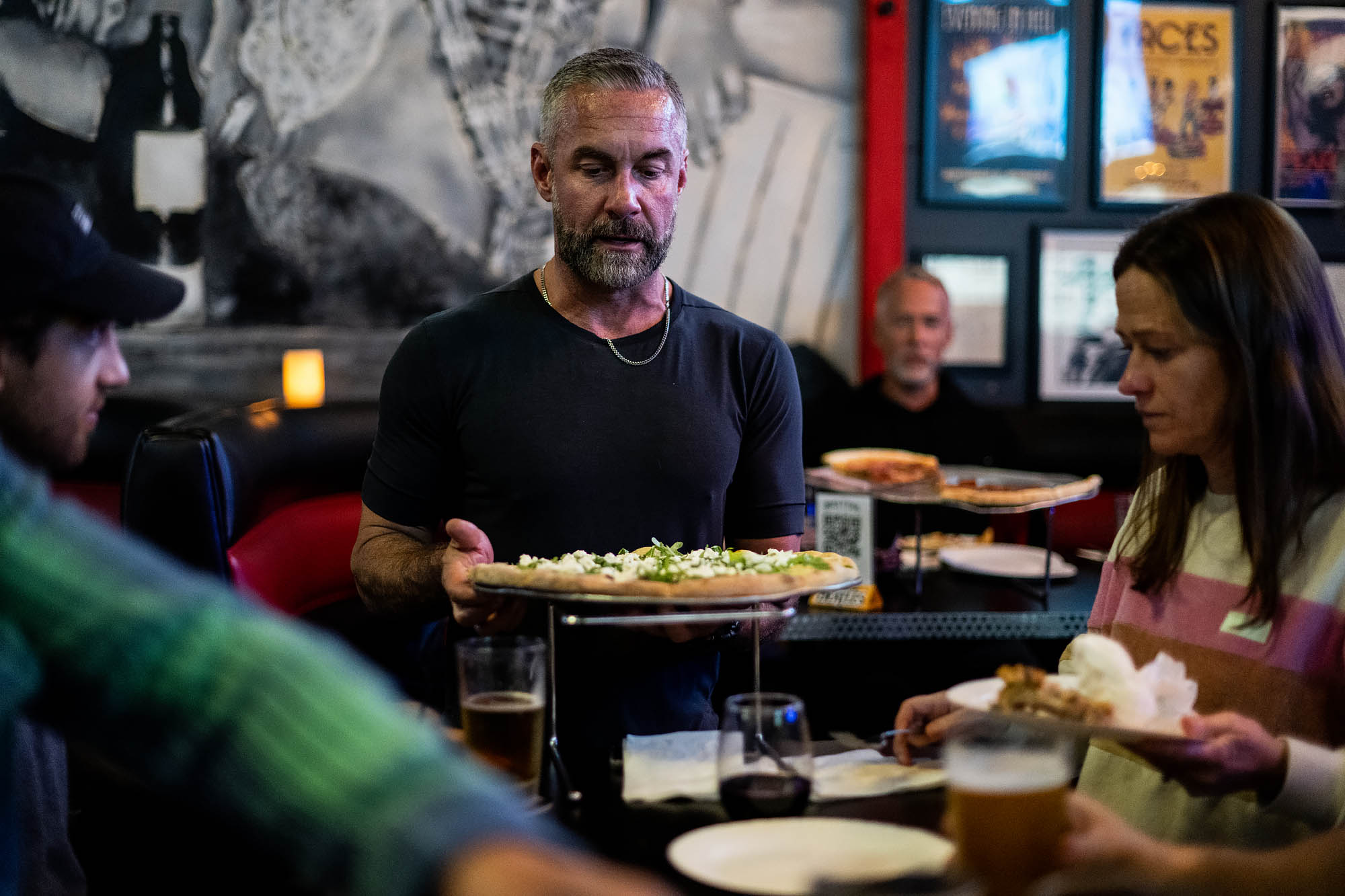 A man with silver hair and a dark t-shirt places a pizza on a table at a dark restaurant.