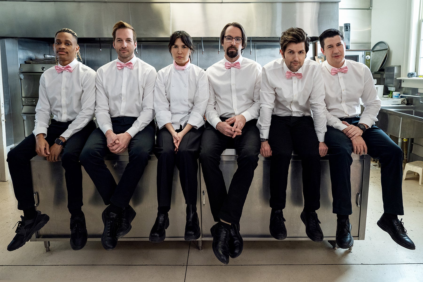 Six caterers in white shirts, pink bow ties, and black pants sit across a counter.