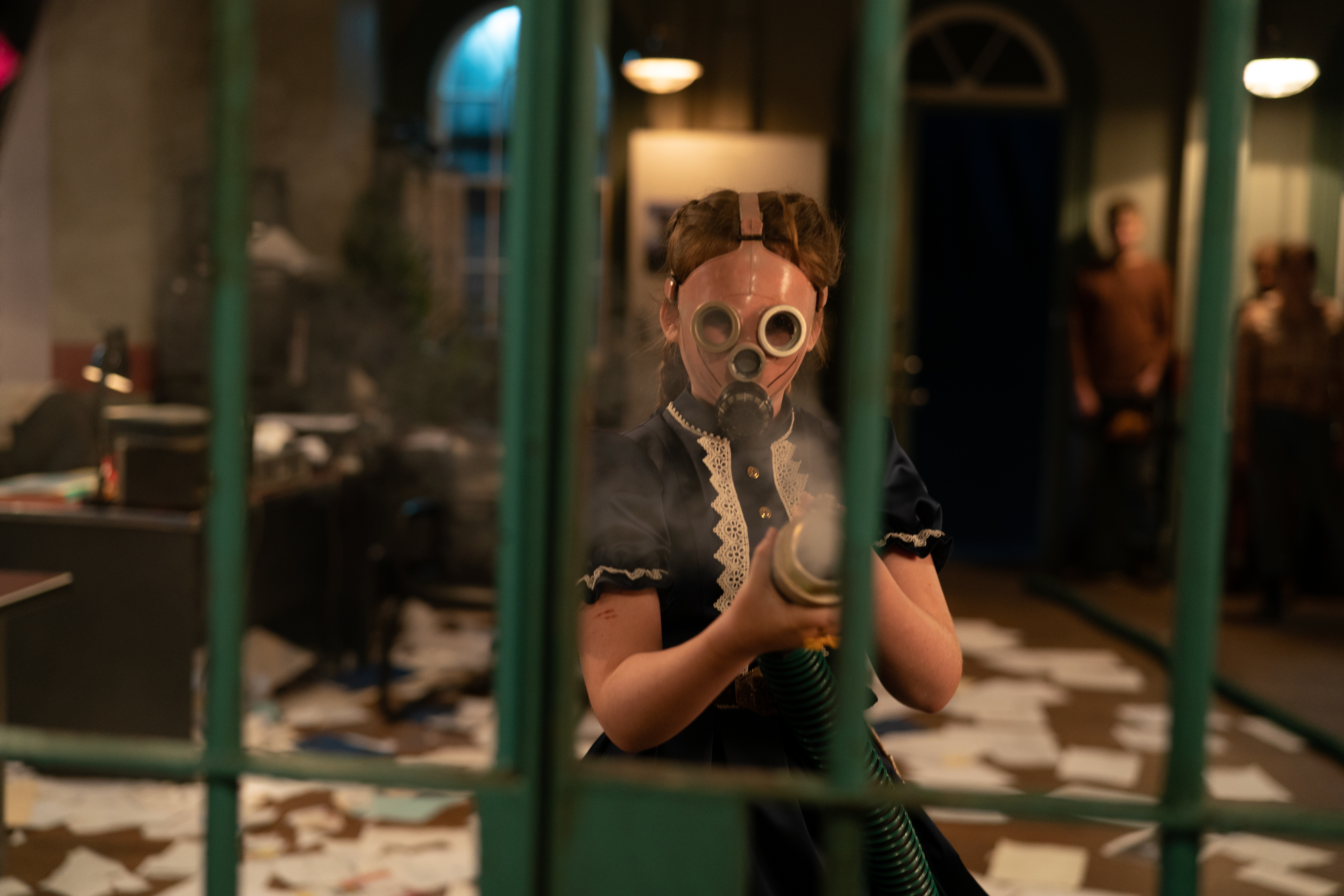 A young girl in a Puritan dress and a creepy goggle-eyed gas mask sprays gas through the green painted bars of a jail cell in the 2023 Children of the Corn