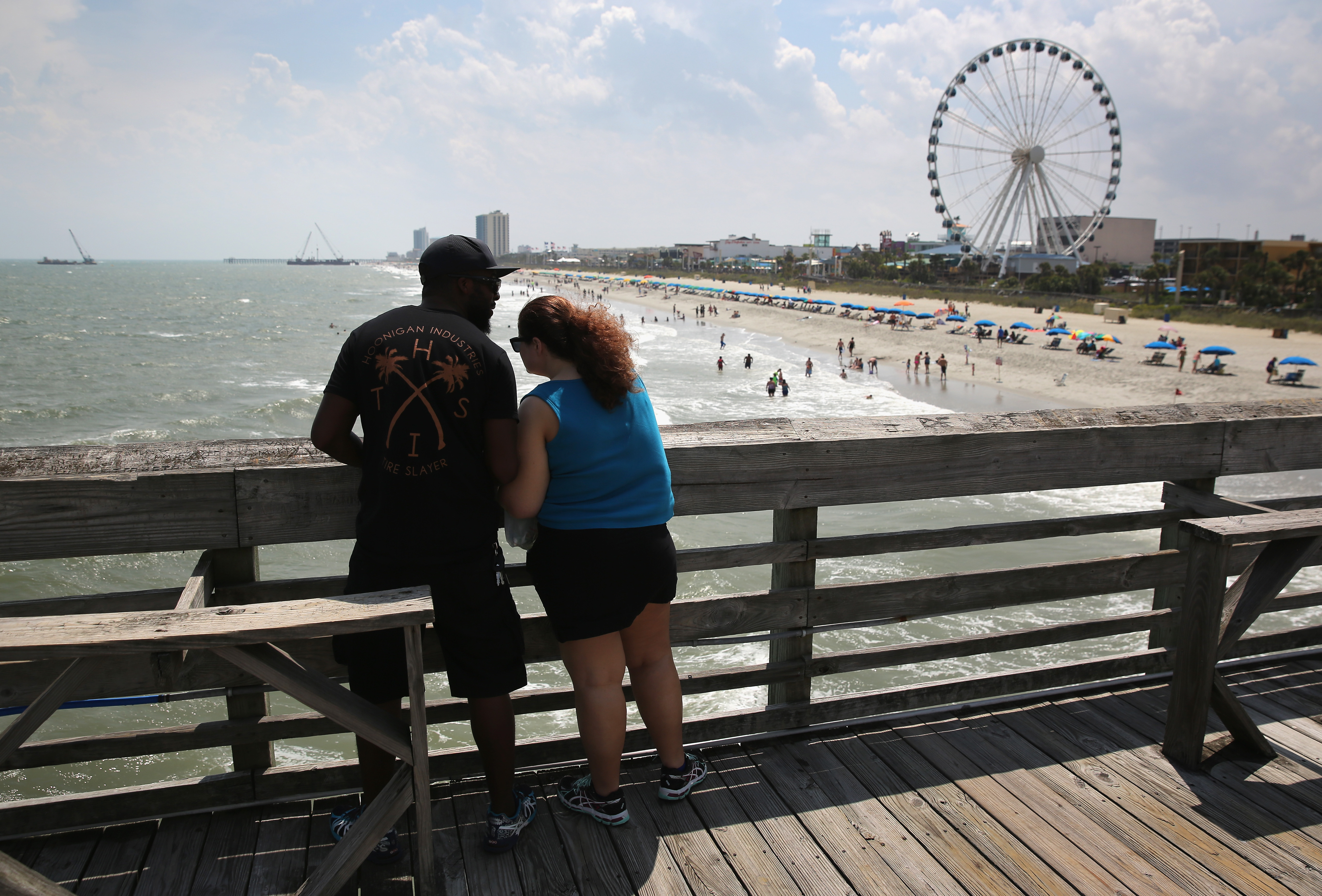 A couple stands on a pier overlooking the beach and the SkyWheel on July 13, 2015 in Myrtle Beach, South Carolina. The 187 foot tall ferris wheel, built in 2011, is the biggest east of the Mississippi River and dominates the skyline of the tourist beach destination. The “Grand Strand” beach stretches almost uninterupted for more than 60 miles of Atlantic coastline and is Myrtle Beach’s top attraction, bringing in millions of tourists each year.
